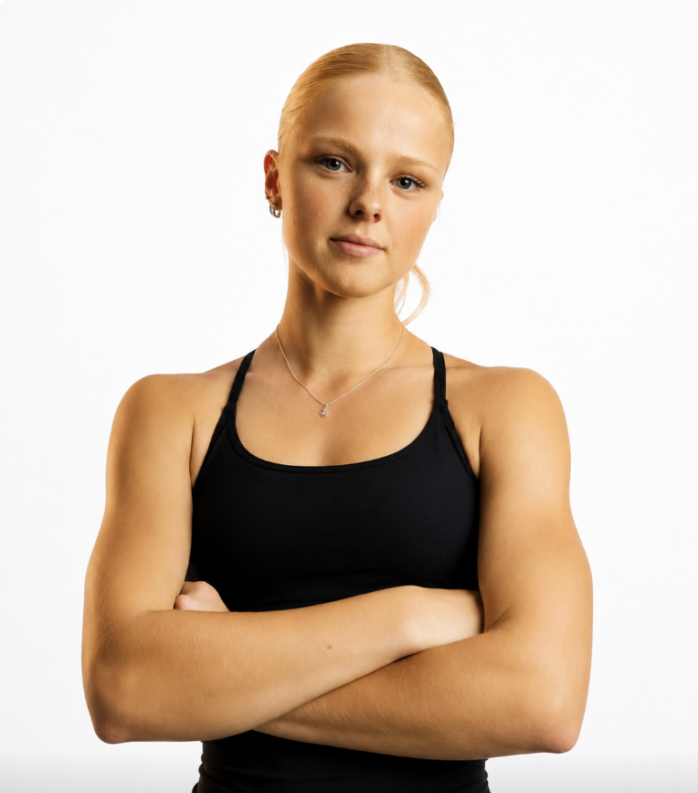 A young woman with blonde hair tied back, wearing a black sports bra, silver jewelry, and standing with arms crossed against a plain white background.