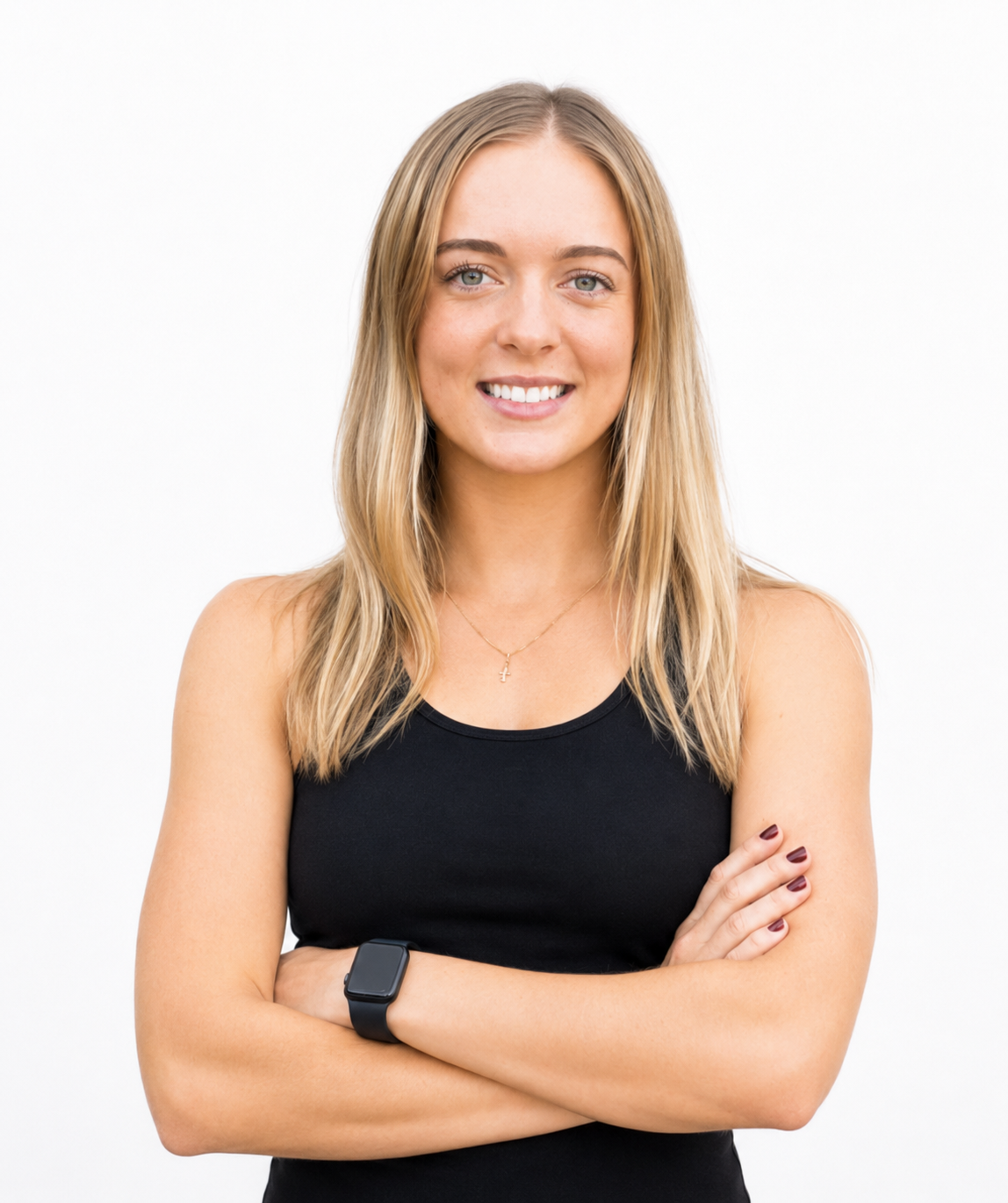 A young woman with blonde hair, wearing a black sleeveless top, smiling with arms crossed, standing against a plain white background.