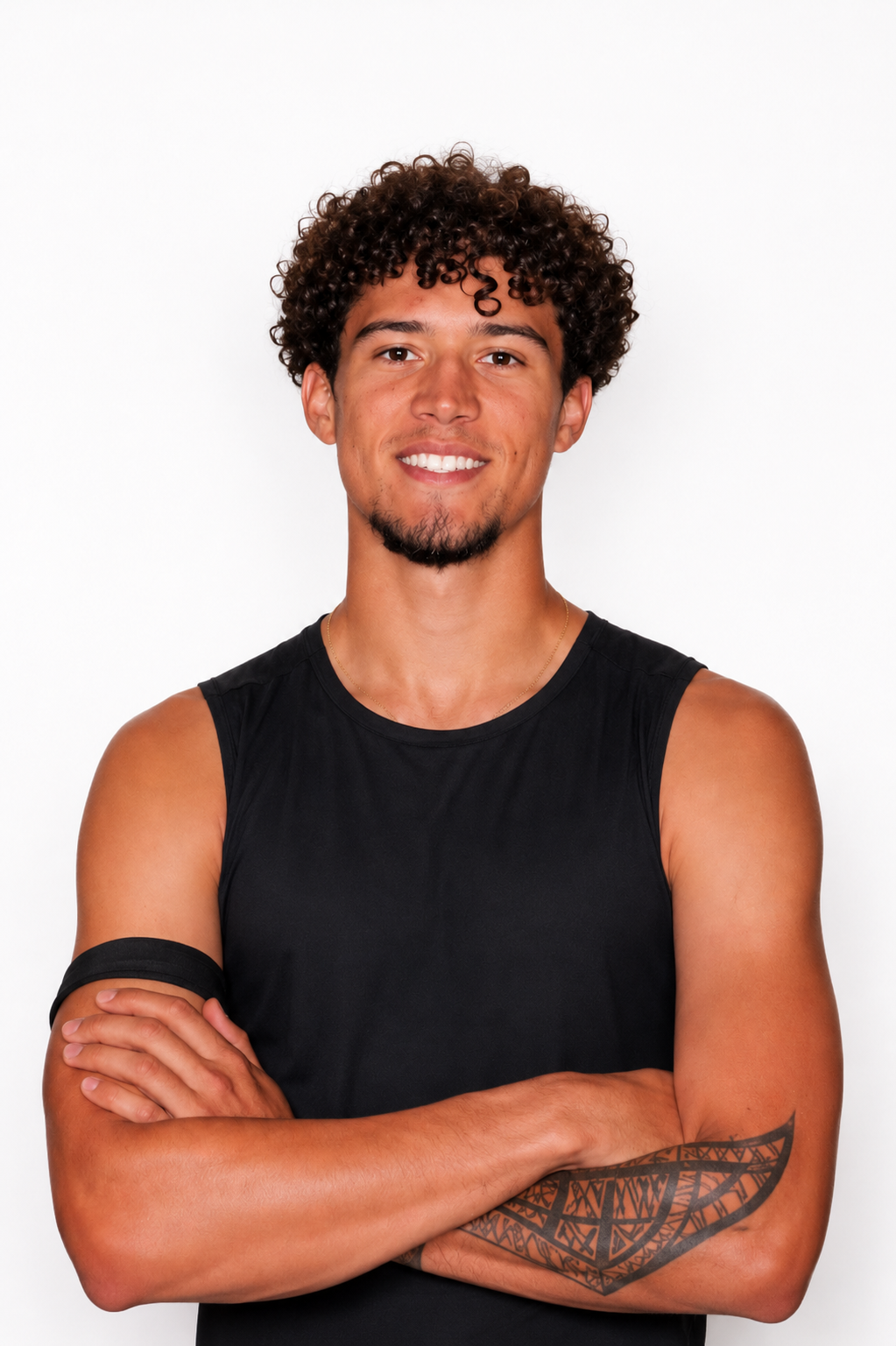 Portrait of a young man with curly hair wearing a black sleeveless shirt, standing with arms crossed against a white background.
