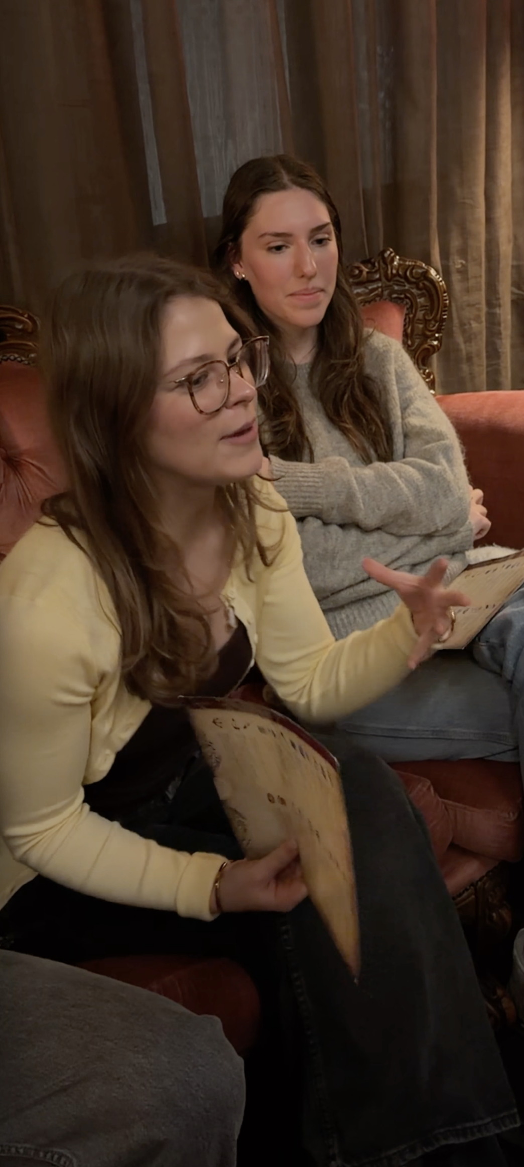 Two women sitting on a vintage upholstered sofa in a cozy wooden-paneled room, engaged in conversation, one holding a menu or a brochure.