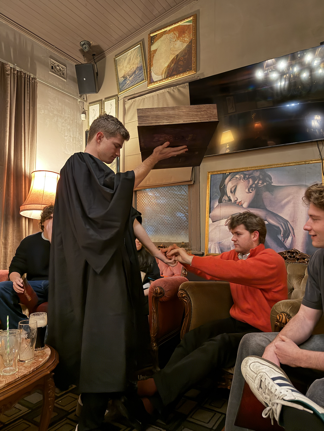 Young man in black robes kneels and holds hands with another young man in a red sweater, who is sitting on a vintage sofa in a cozy, dimly lit room decorated with paintings and a large mirror.