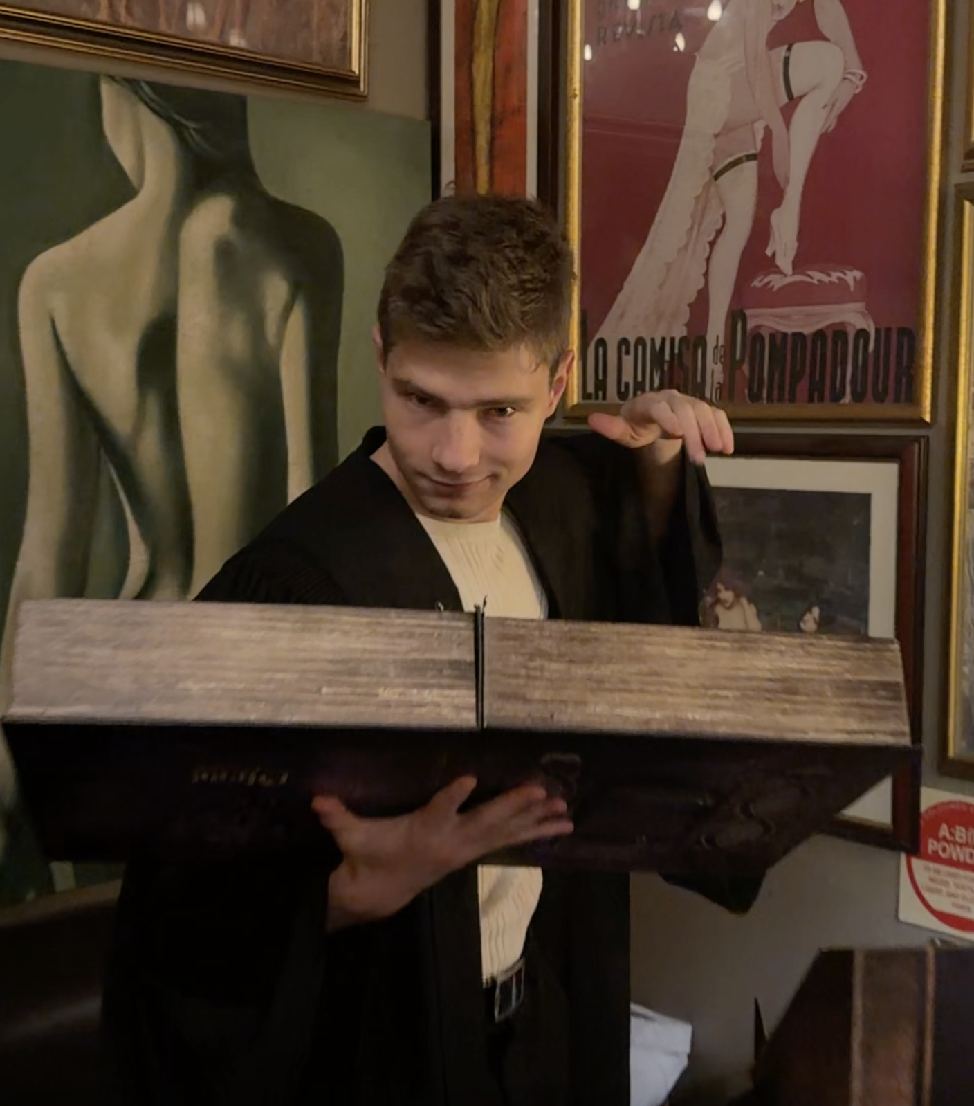 A young man with short brown hair holding a large book in front of a wall decorated with vintage posters,