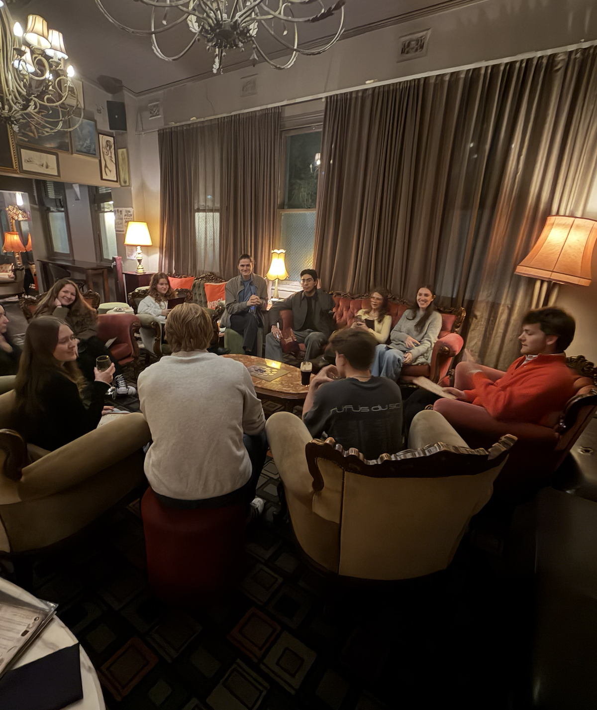 Group of people sitting on sofas and chairs in a cozy, warmly lit living room, socializing and drinking.