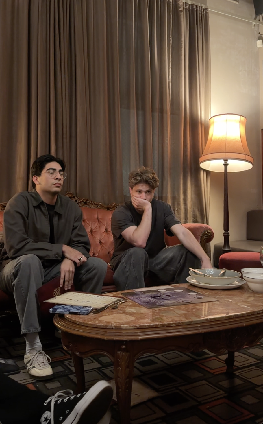 Two young men sitting on a red velvet sofa in a dimly lit room with curtains and a standing lamp. One appears bored or asleep, and the other covers his mouth with his hand, looking surprised or concerned. A table in front has bowls, a plate, and some game pieces.