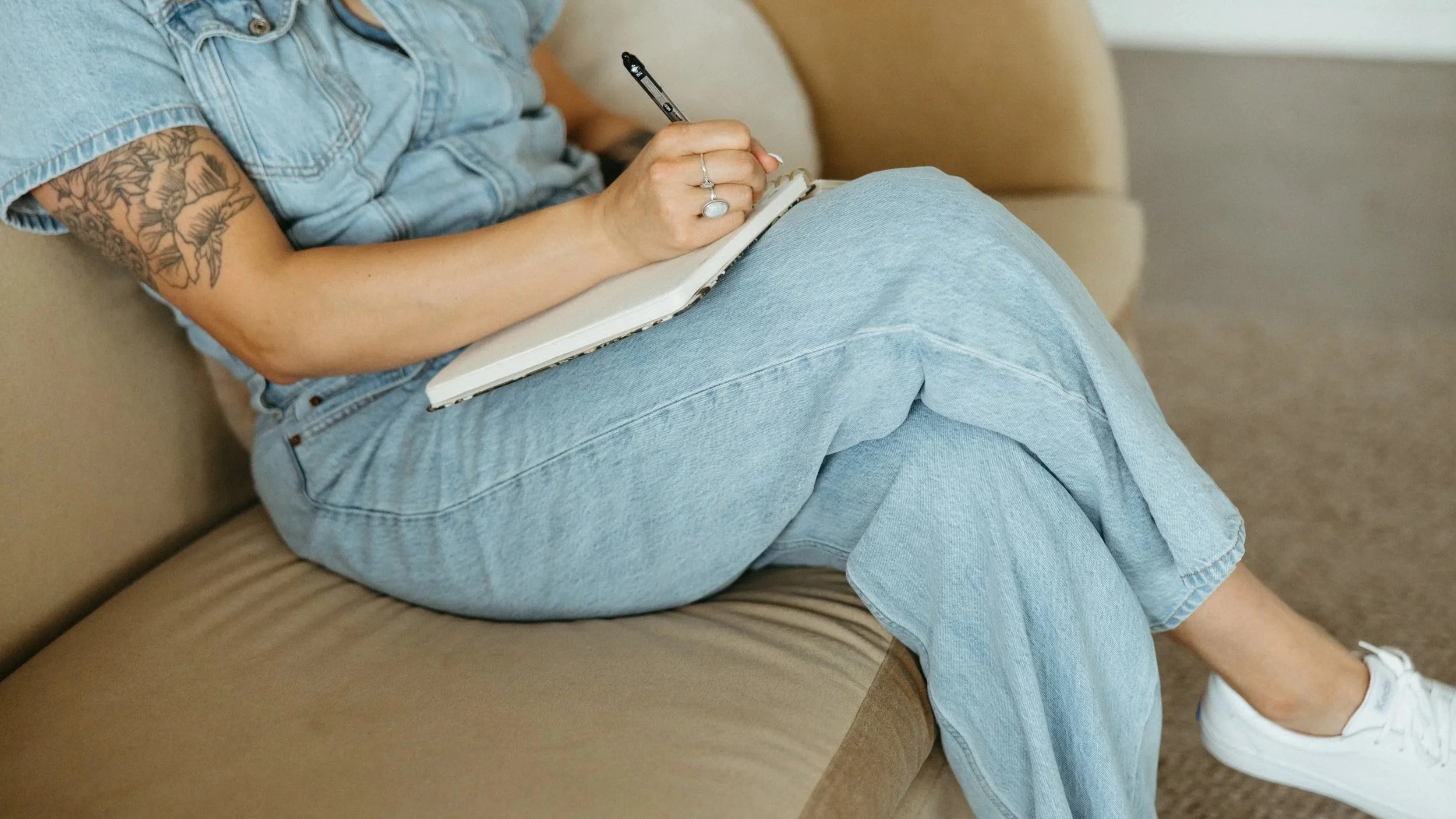 A person sitting on a beige couch, wearing a denim shirt and jeans, holding a smartphone in their right hand, and writing or drawing in a notebook resting on their lap. The person has a large floral tattoo on their left arm.