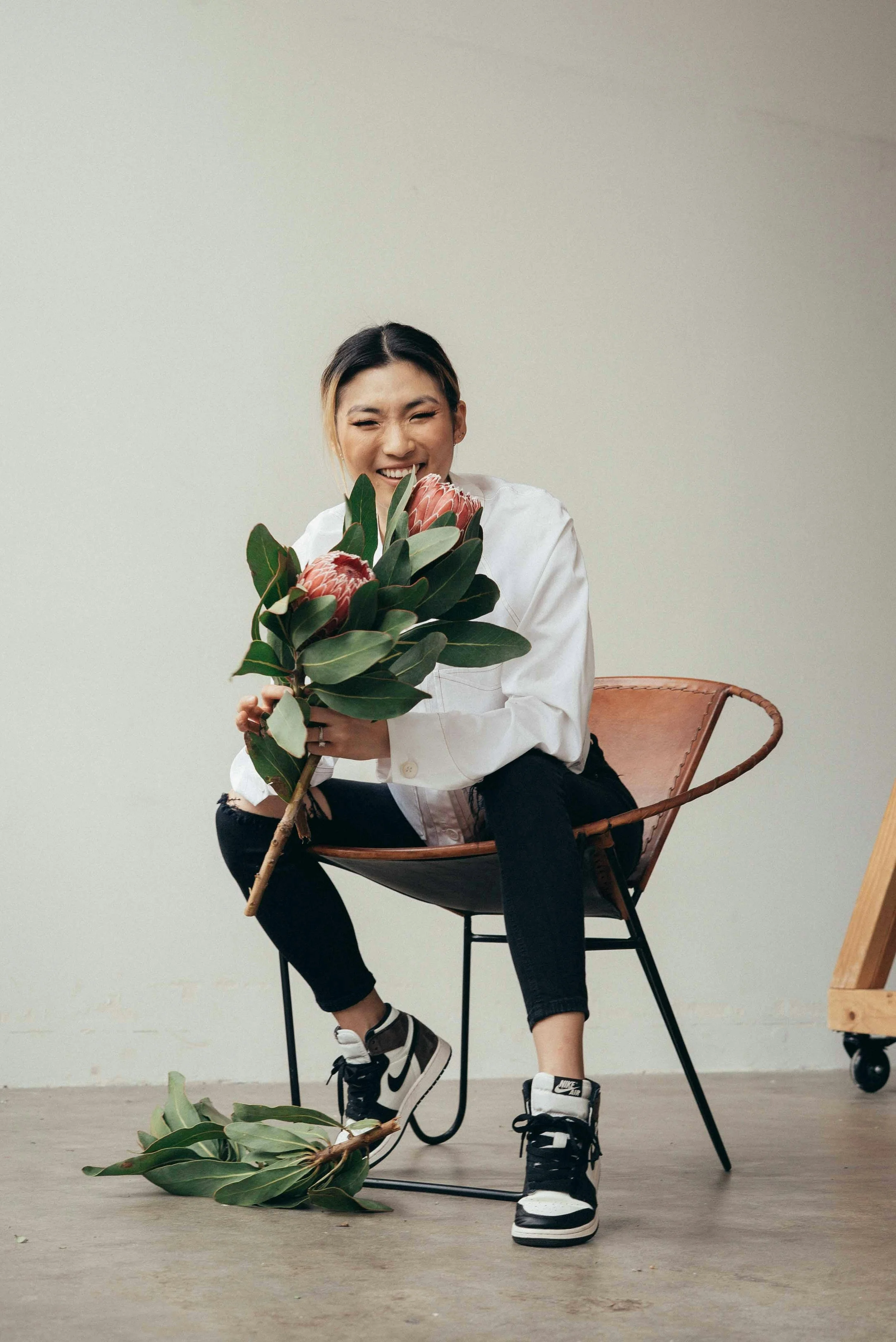 A young woman with dark hair tied in a ponytail smiling and winking while sitting on a modern chair in a minimalist room. She is holding a large bouquet of tropical flowers and greenery. She is dressed in a white shirt, black ripped jeans, and Nike sneakers. There are some large leaves from the bouquet on the floor and a wooden piece of furniture on wheels is partially visible on the right.