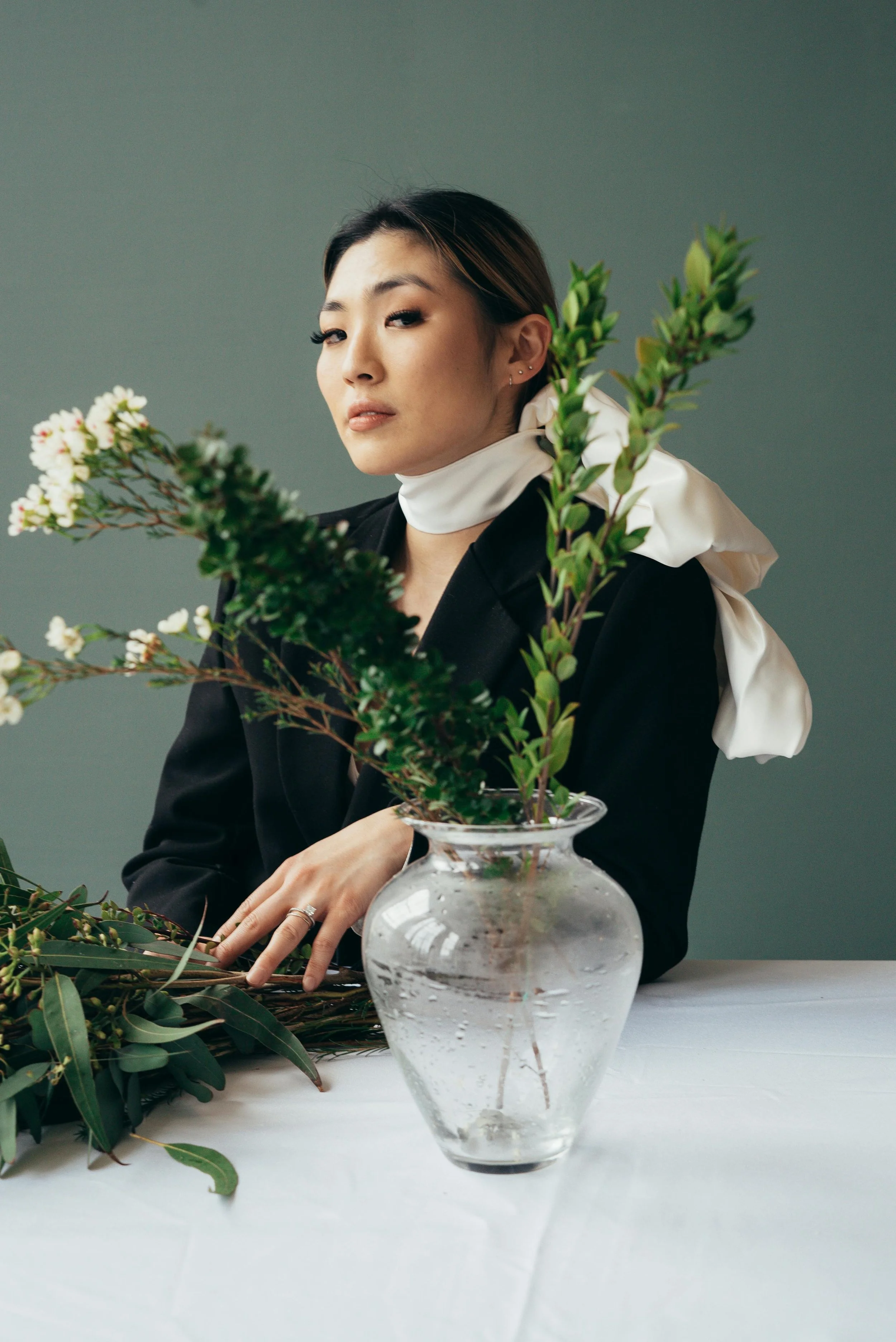 A woman with short dark hair and earrings, wearing a black blazer and white scarf, looks at the camera while arranging greenery and flowers in a clear glass vase on a white table against a gray background.