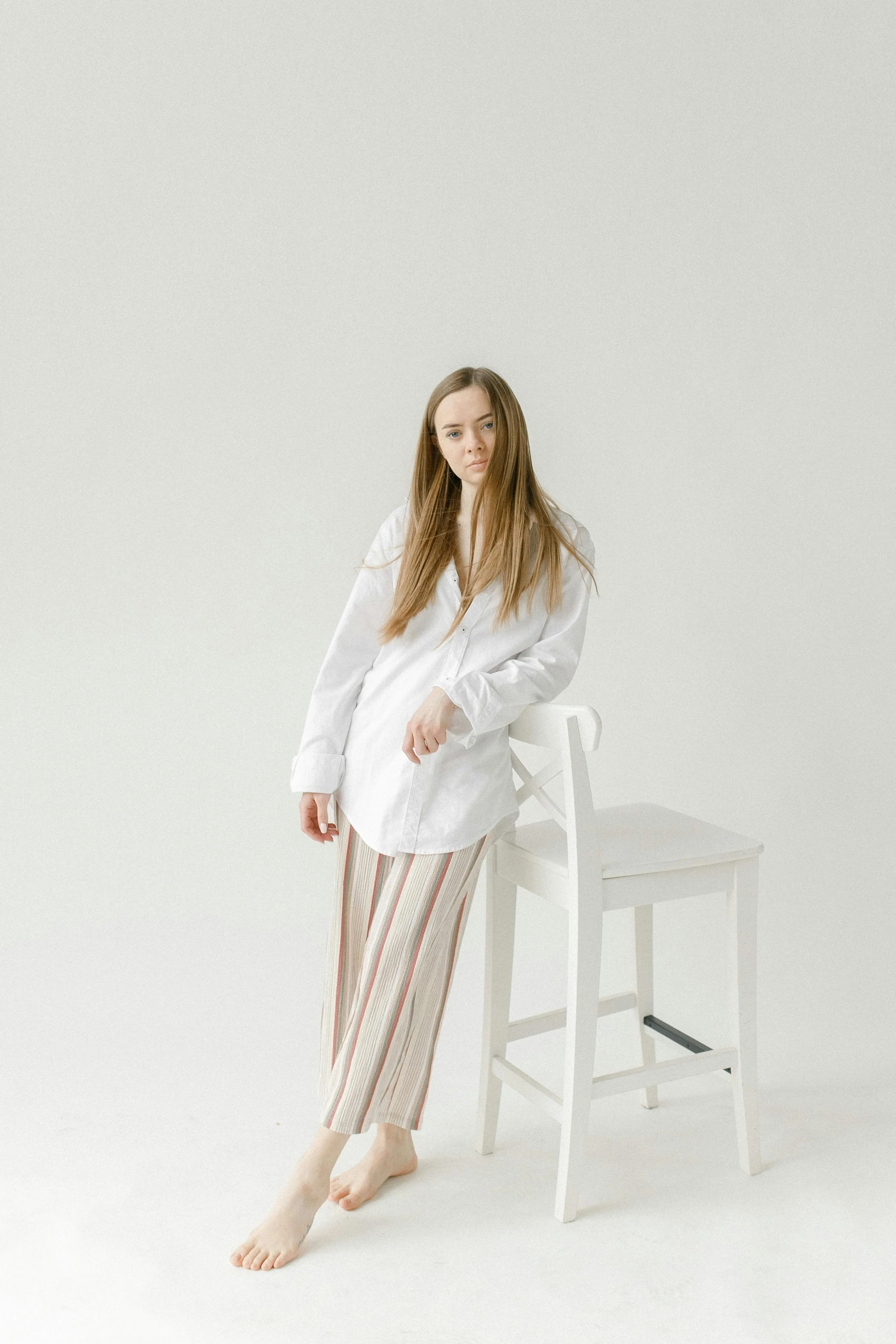 A young woman with long, straight, light brown hair and fair skin standing barefoot next to a white chair against a plain, light-colored background. She is wearing a loose white shirt and striped pants in pastel colors.