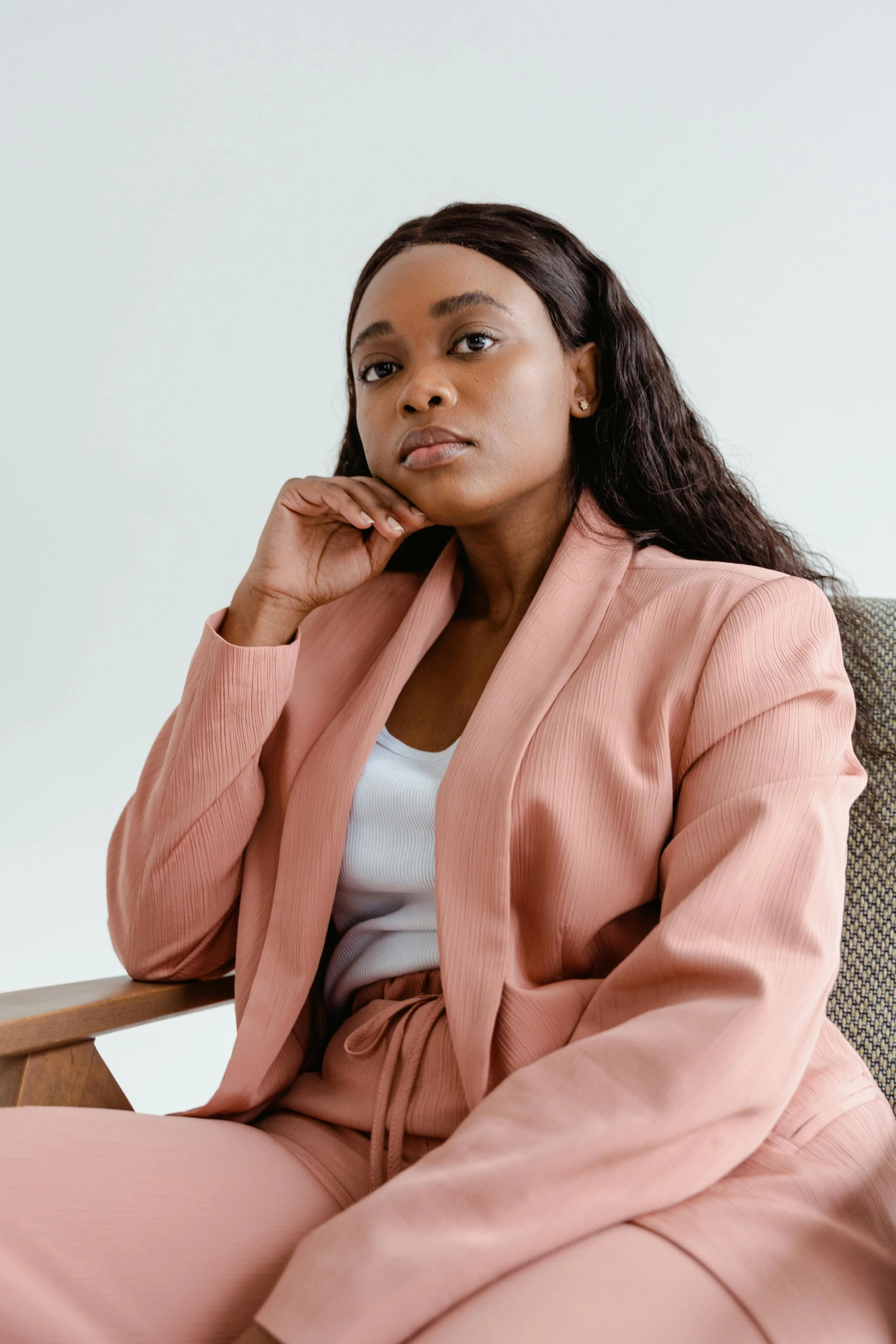 A woman in a pink tracksuit sitting in a chair with her hand resting on her chin, looking into the distance with a neutral expression.