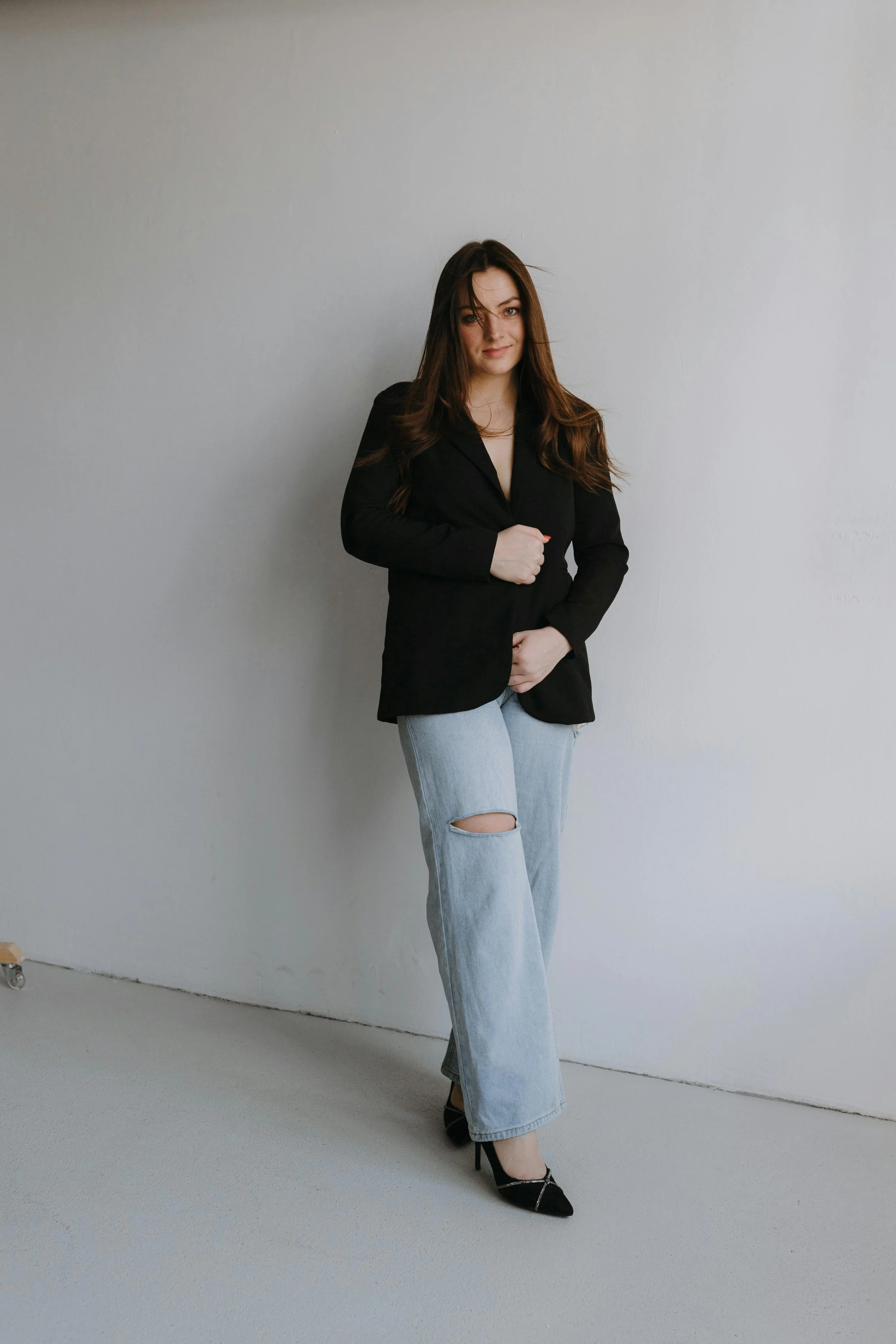A woman with long brown hair, wearing a black blazer, light blue ripped jeans, and black high heels, standing against a plain white wall.