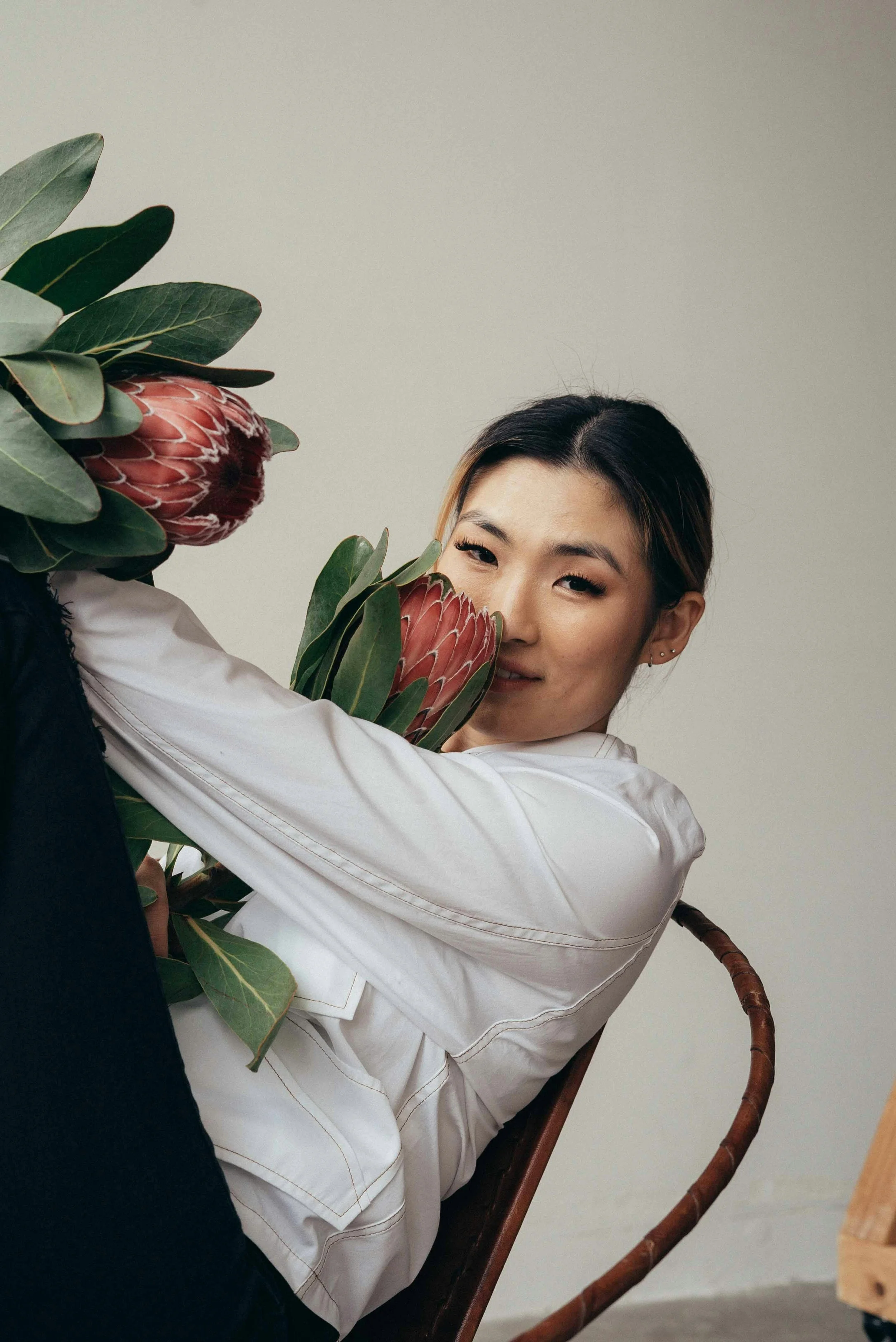A woman sitting on a wooden chair, leaning on large green leaves and protea flowers, with her arm resting on the plant, wearing a white shirt and earrings, against a neutral background.