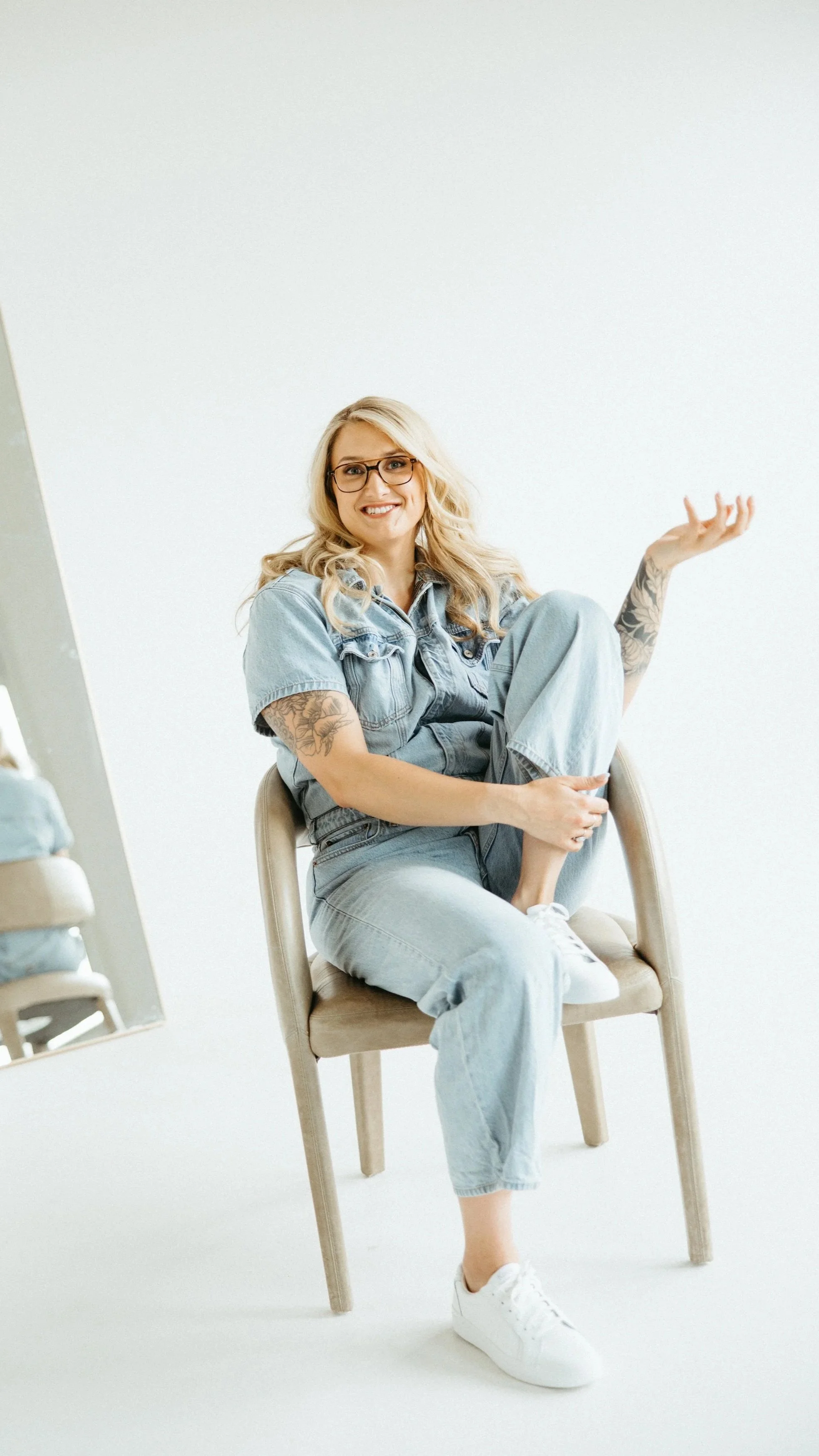 A young woman with blonde hair, glasses, and tattoos, sitting on a beige chair against a white background, smiling, with her right arm raised and her left foot over her right knee.