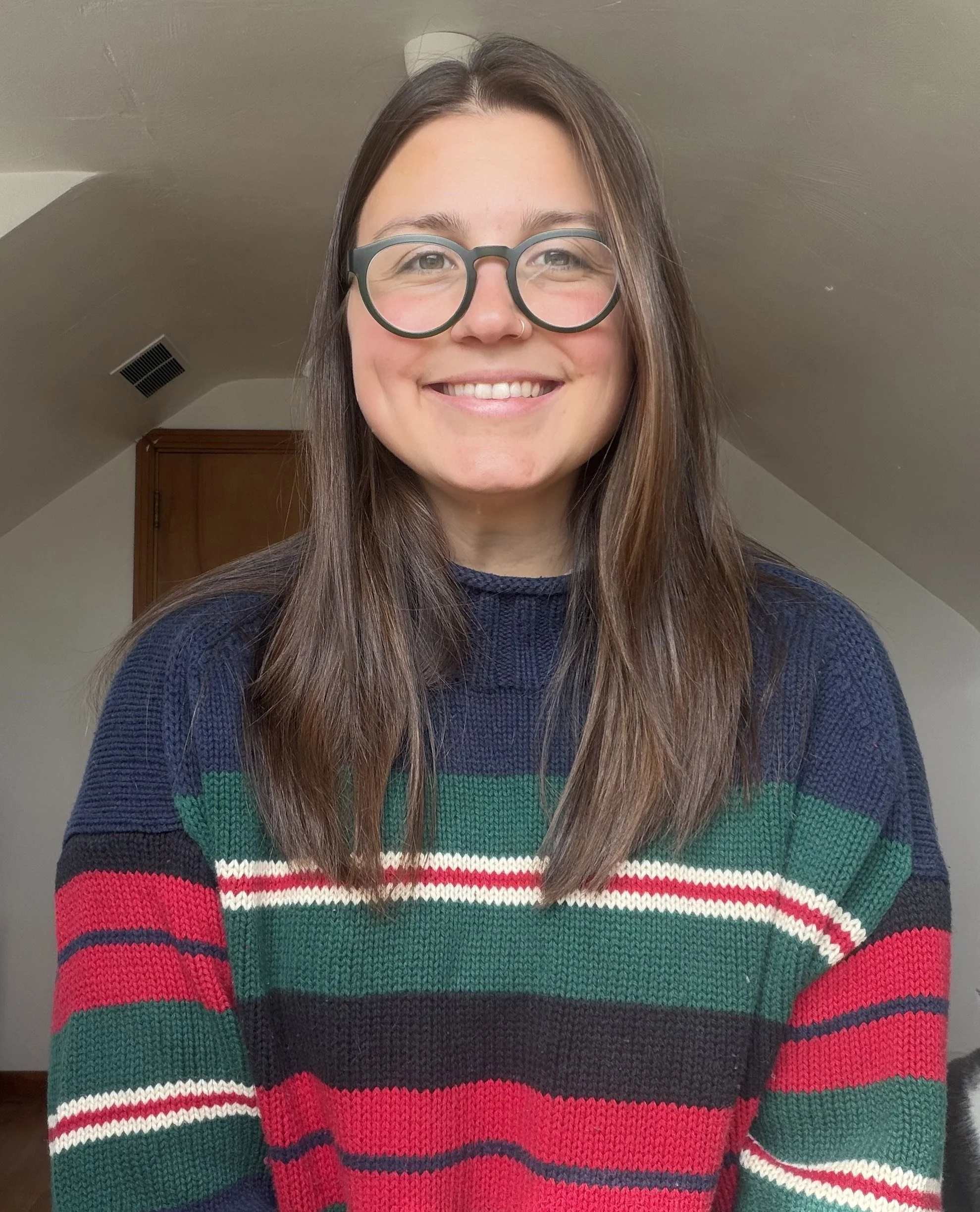 A smiling woman with long brown hair, wearing glasses and a rainbow-colored striped sweater, standing in a room with a beige ceiling and light fixture.