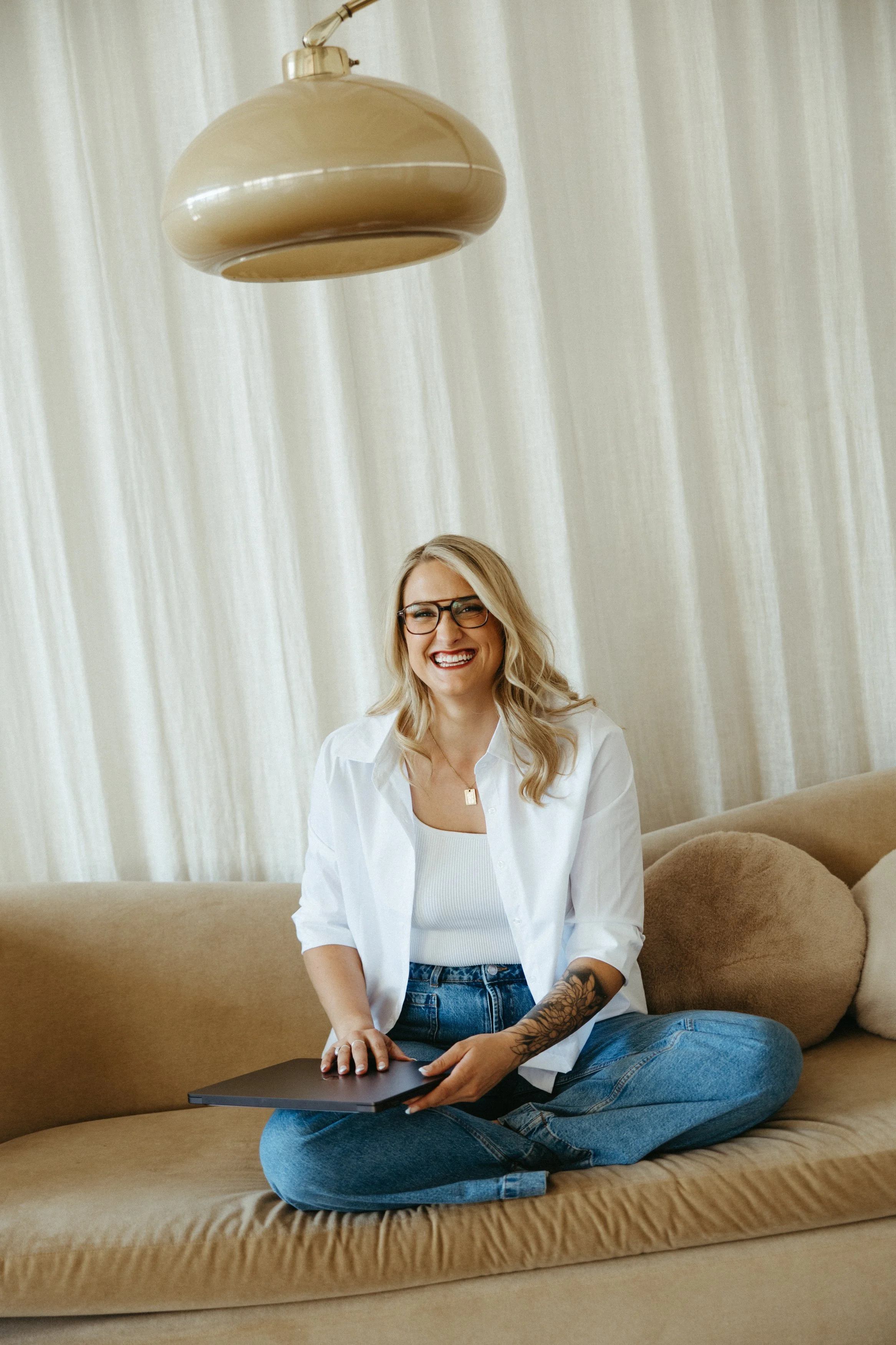 A woman with glasses, blonde hair, and a tattooed arm, sitting cross-legged on a beige sofa, holding a closed laptop and smiling, in front of cream-colored curtains.