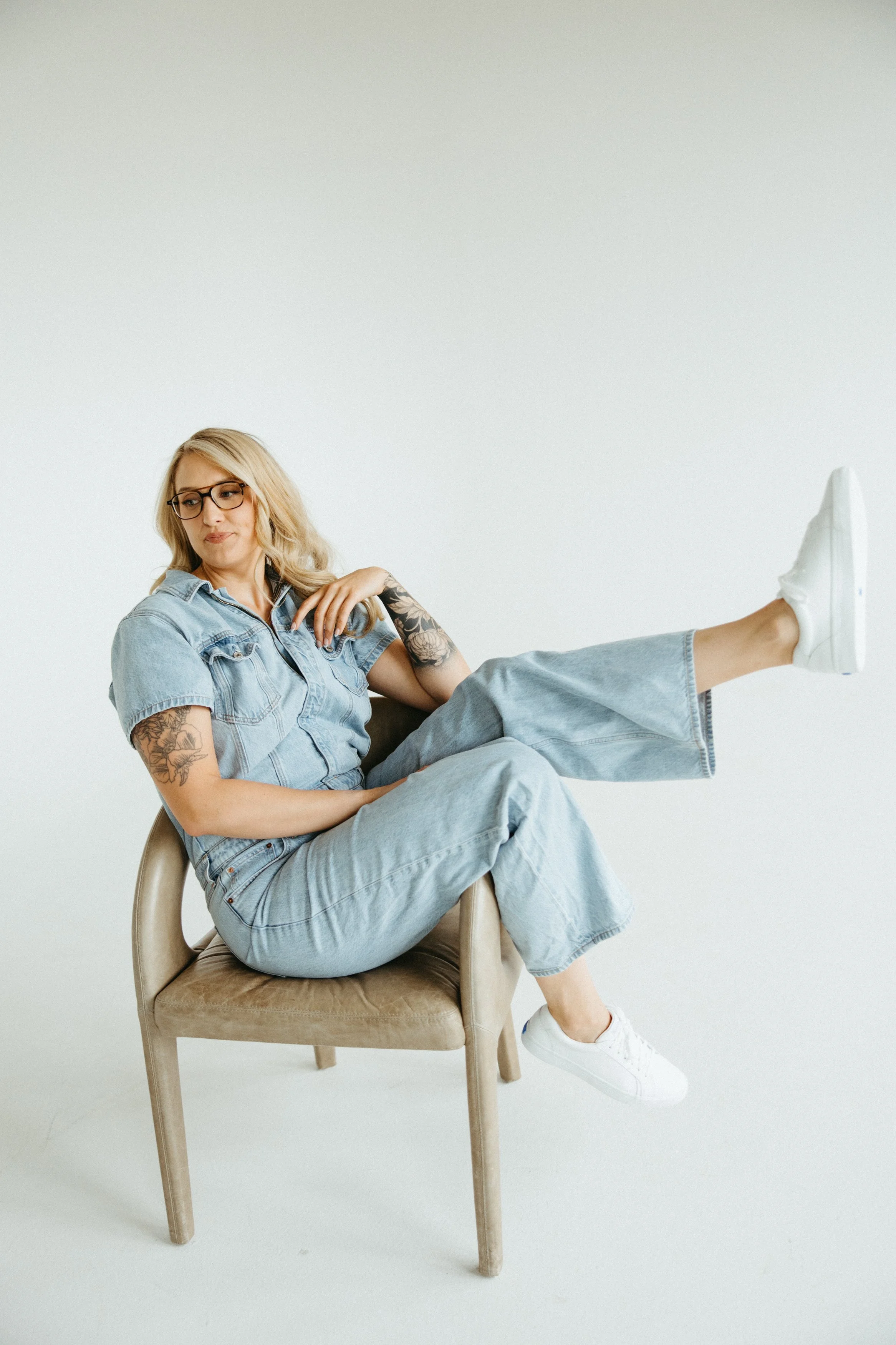 A woman with blonde hair, glasses, tattoos on her arms, wearing a denim jacket and jeans, sitting on a beige chair with one leg raised and wearing white sneakers, against a plain white background.
