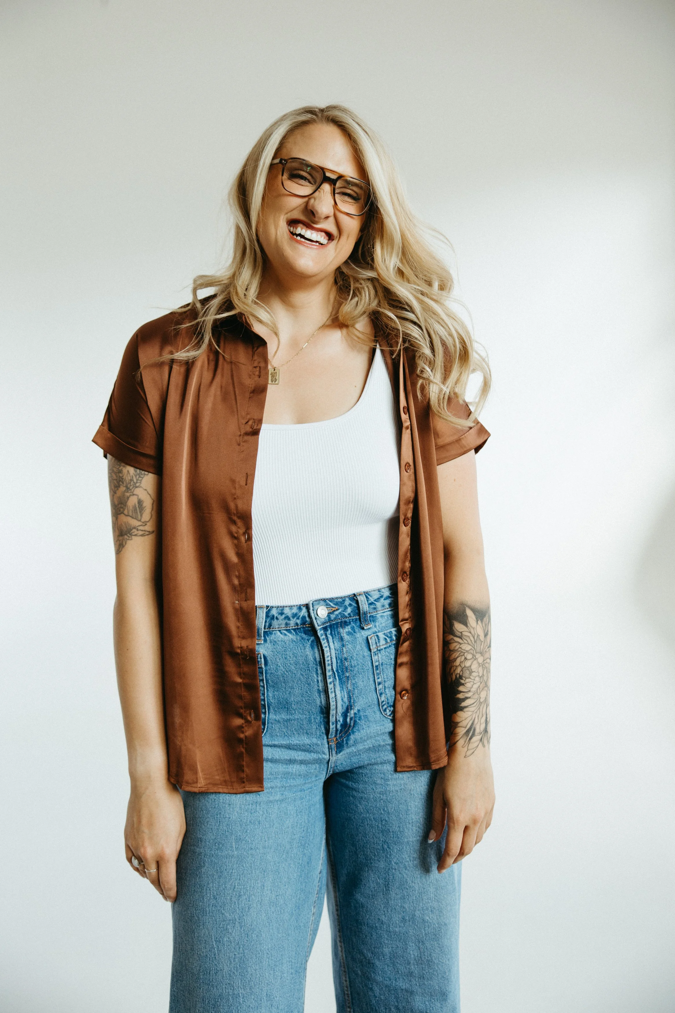 A woman with blonde hair and glasses, smiling and wearing a white tank top, a brown satin shirt, blue jeans, and has tattoos on her left arm, standing against a plain white background.