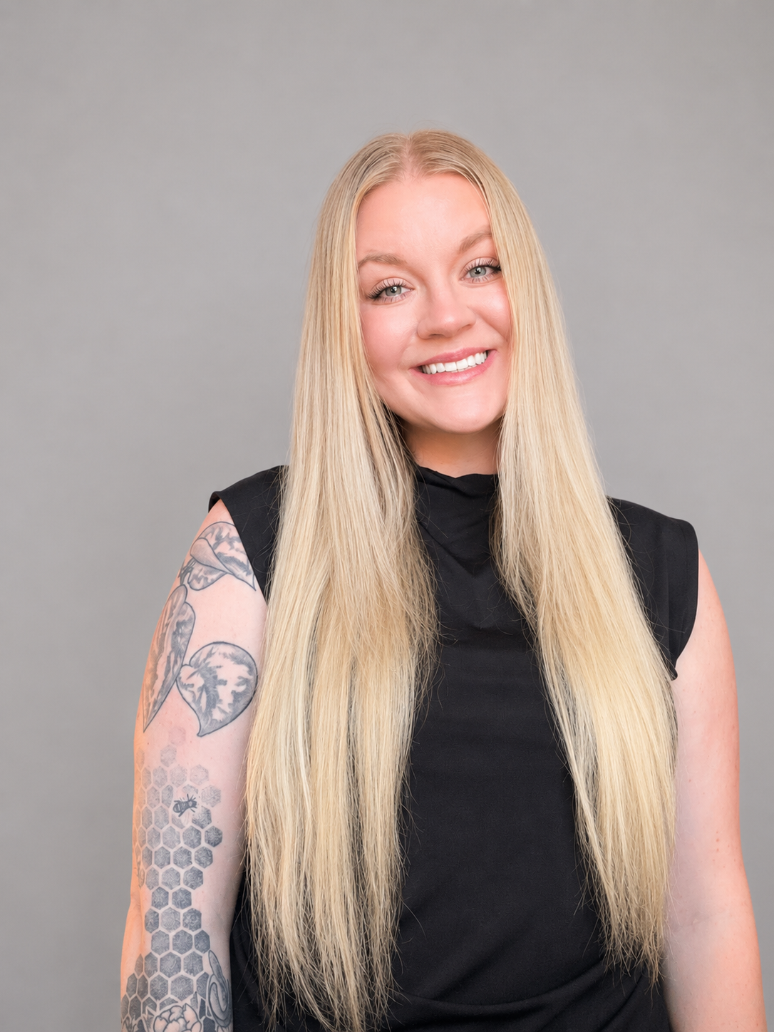 A young woman with long blond hair smiling, wearing a black sleeveless top, with a tattooed arm featuring flowers, honeycomb pattern, and an insect, against a plain gray background.
