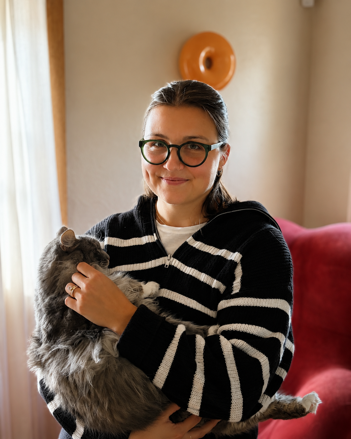 A woman with glasses and a nose ring holding a fluffy grey and white cat indoors near a window and a red couch.