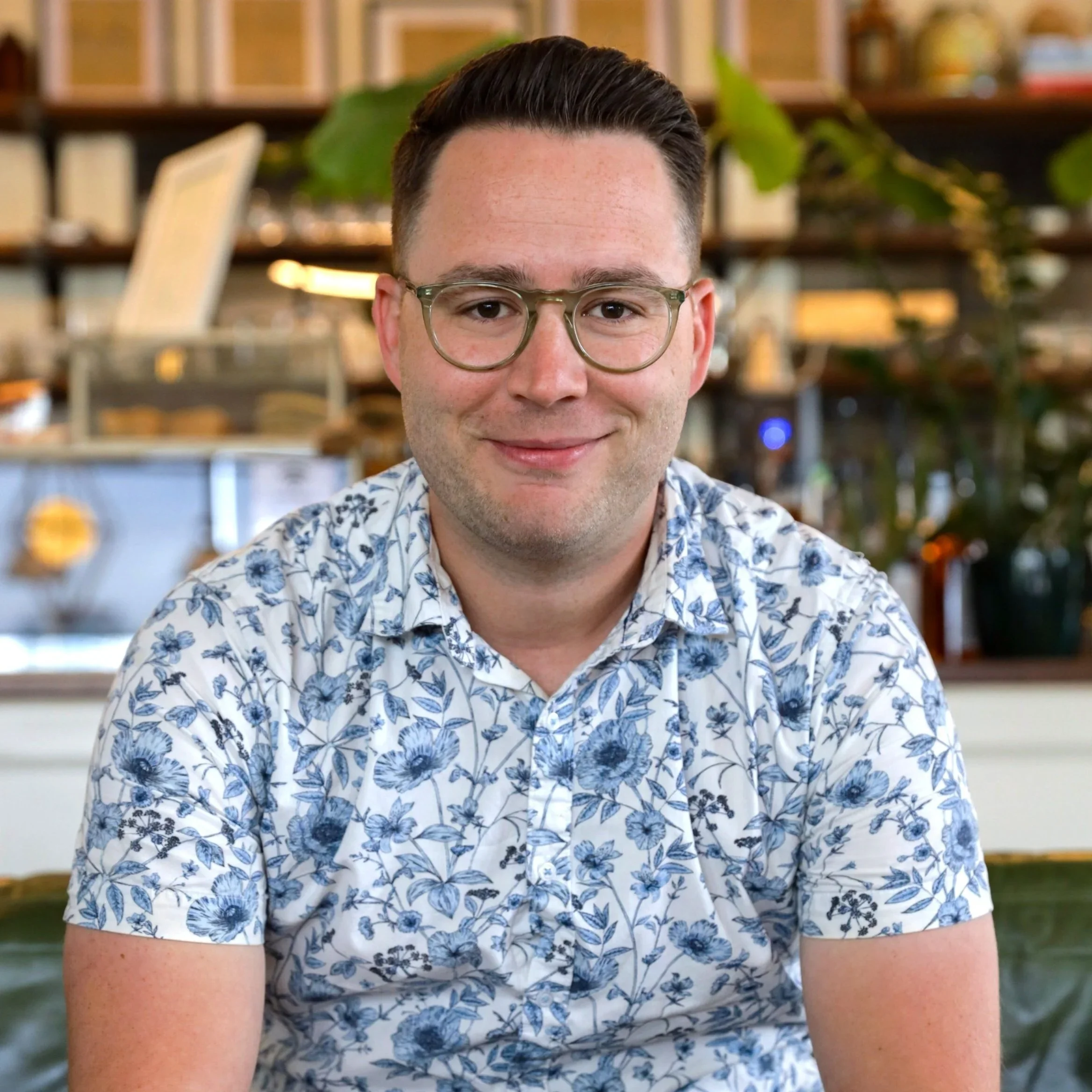Close-up of a young man with dark hair, glasses, and a floral shirt, smiling, with a blurred background of bookshelves and plants in a library or coffee shop.