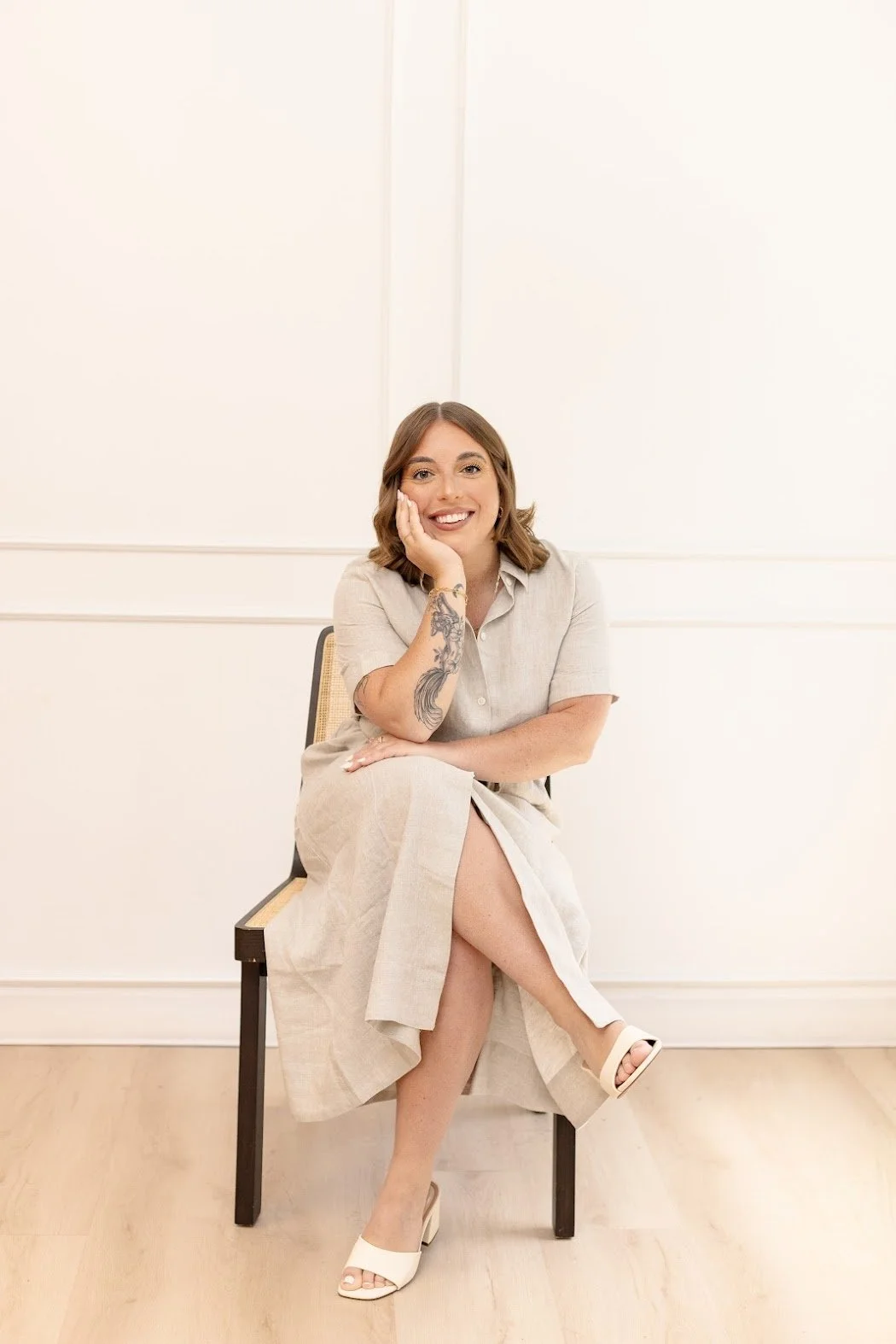 A woman with shoulder-length brown hair, smiling, sitting on a chair with her right elbow resting on her right knee, and her left hand supporting her face. She has a tattoo on her right arm, is wearing a light beige dress, white high-heeled shoes, and is in a room with plain white walls and light wood flooring.