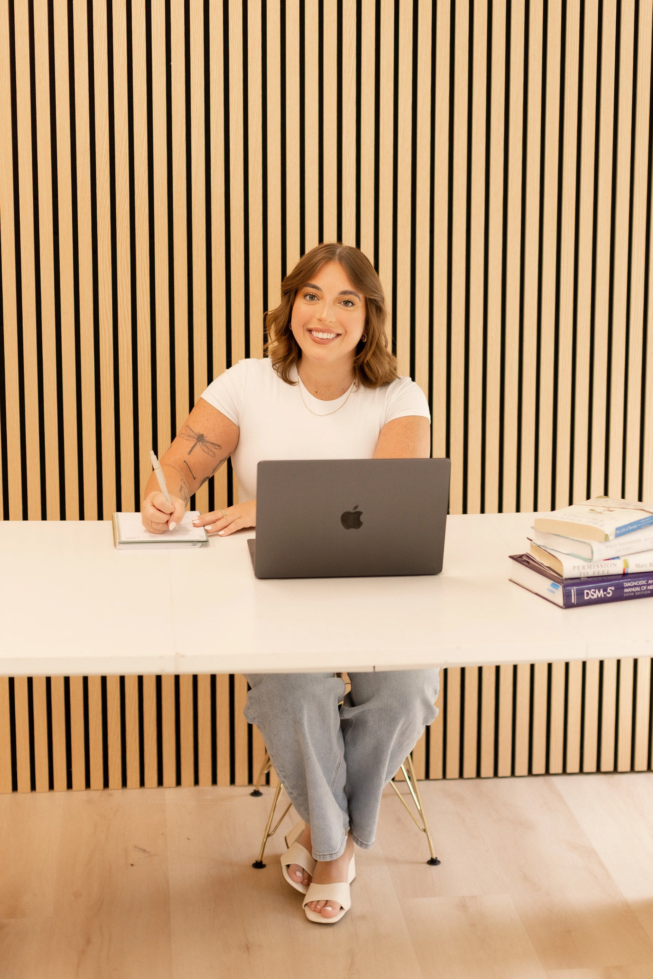 A woman sitting on a chair at a desk, smiling, with a laptop, book, notebook, and papers in front of her. The background features wood paneling.