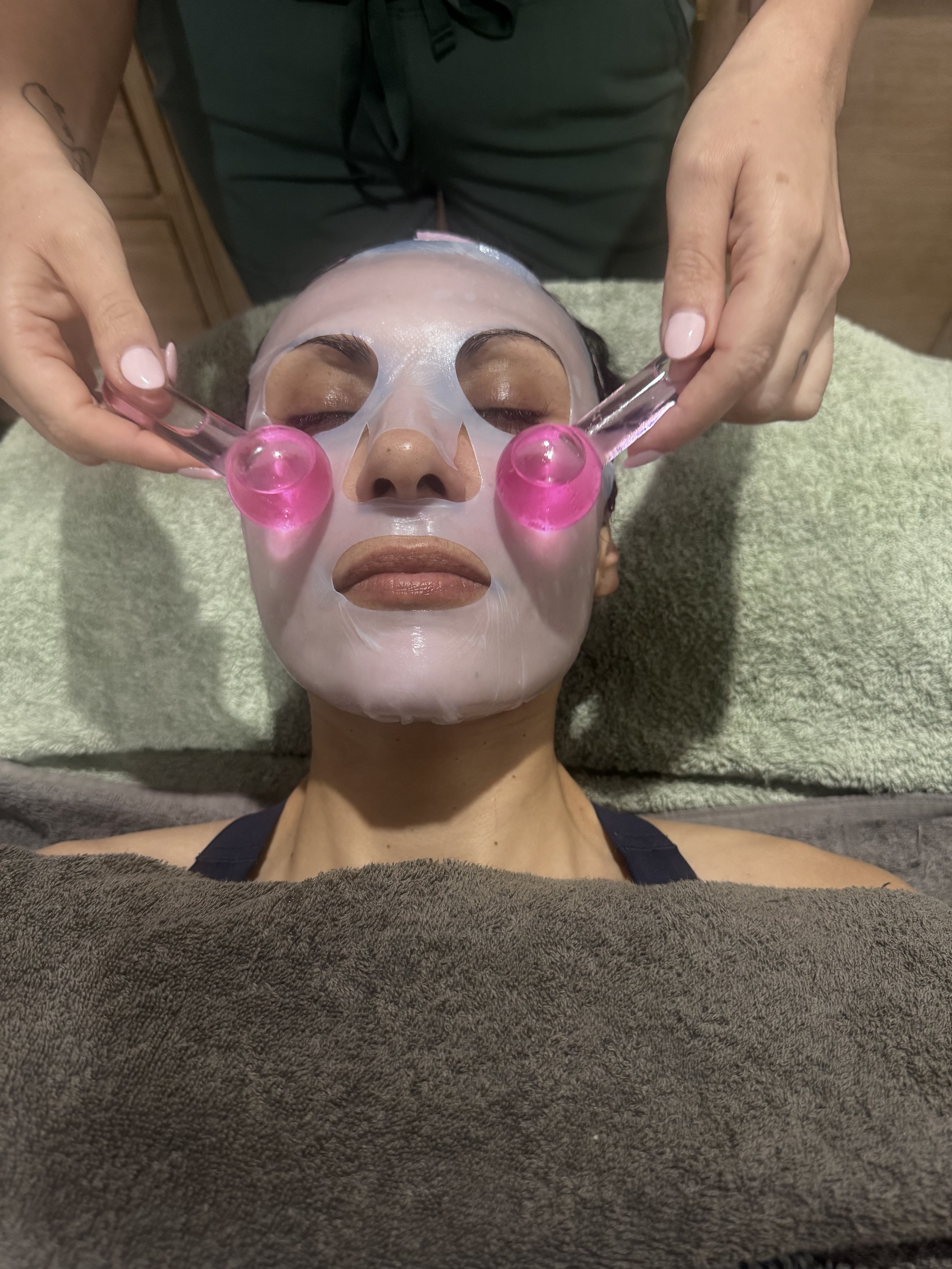 A woman is receiving a facial treatment with a sheet mask and pink facial tools held in her hands, lying on a treatment table covered with a towel.