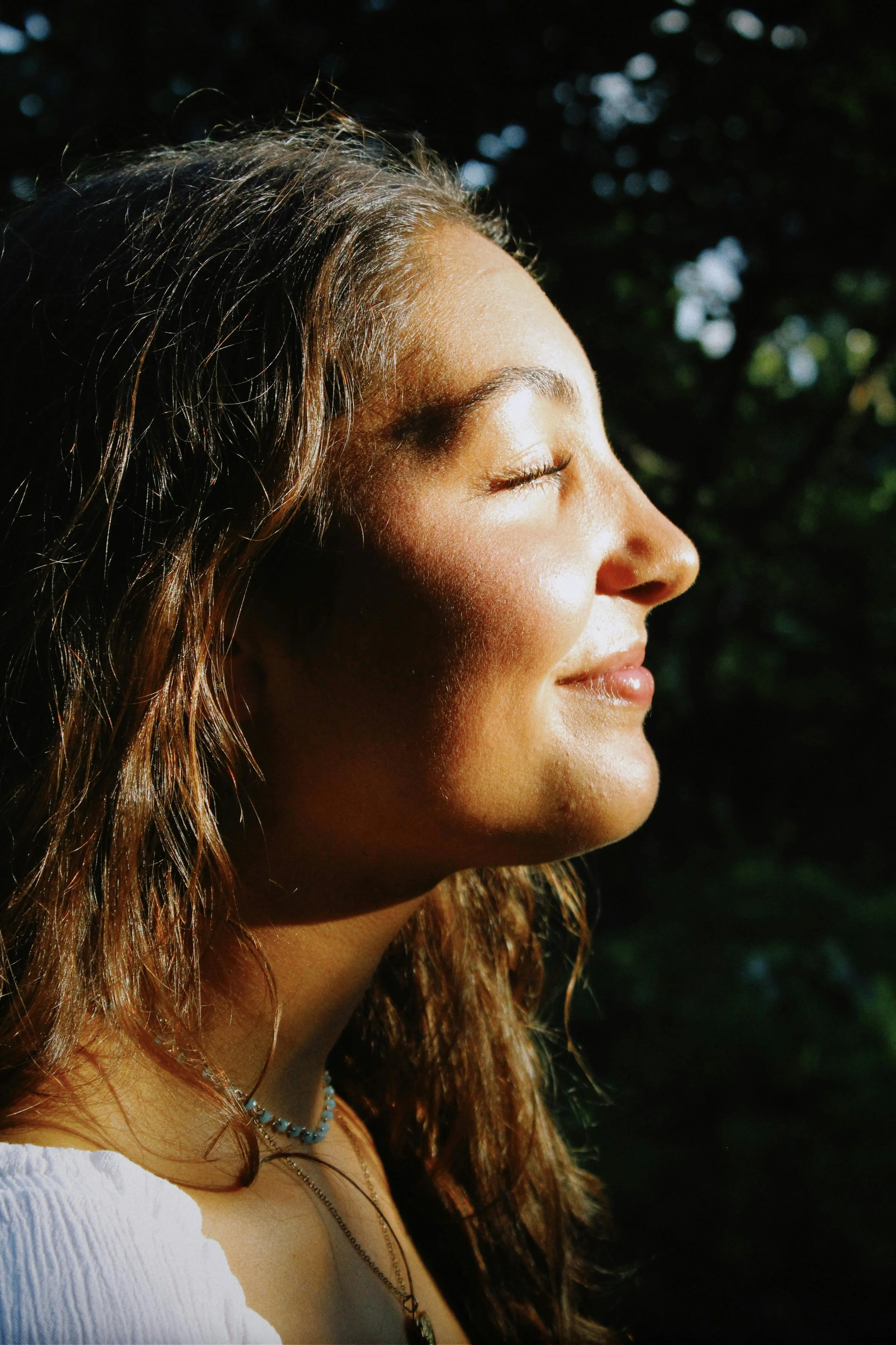 Close-up side profile of a woman with closed eyes, smiling, in sunlight with a blurred dark green background.