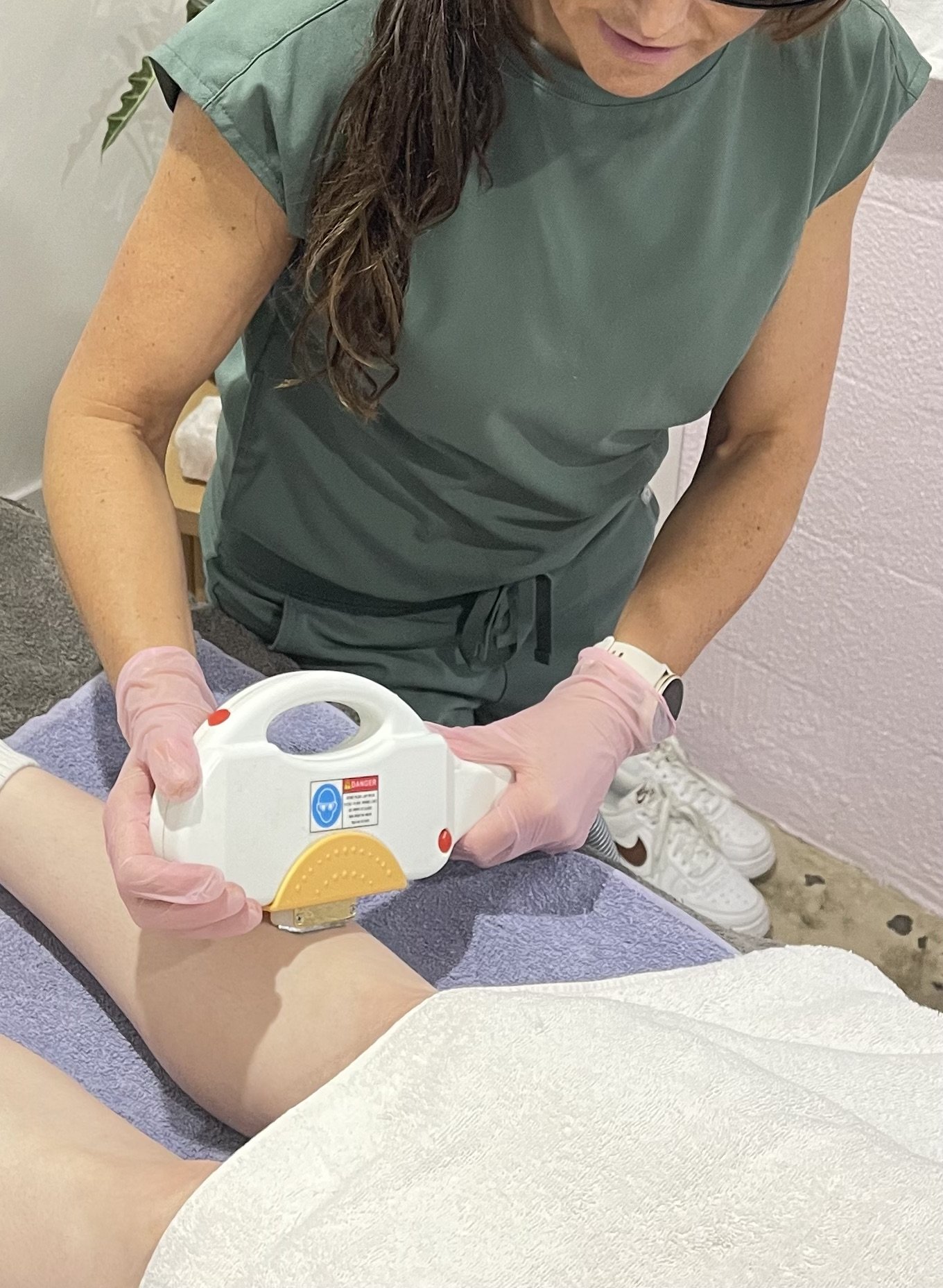 A healthcare worker wearing pink gloves is performing laser therapy on a patient's leg in a clinic with pink walls. The patient is lying on a table covered with a purple towel.