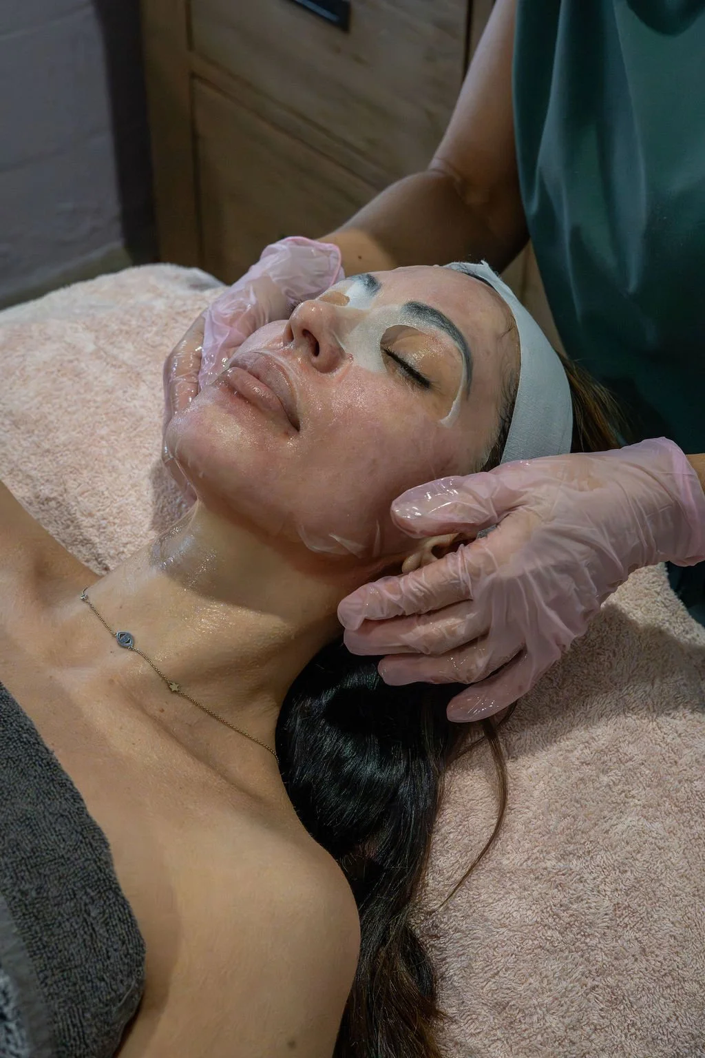 A woman receiving a facial treatment with a mask applied, lying on a spa bed with her eyes closed, wearing a necklace and a headband to hold her hair back.