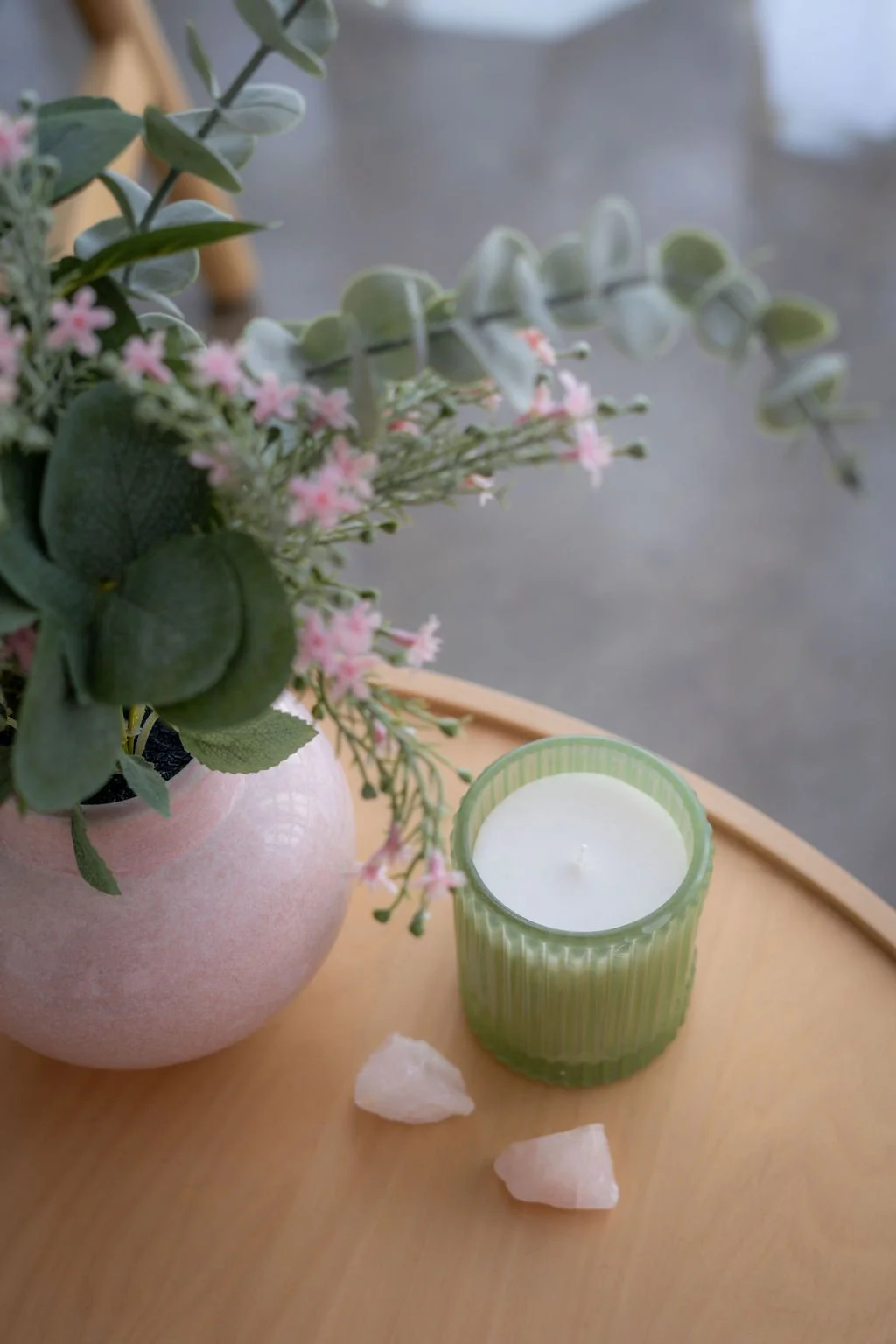A pink vase with green foliage and pink flowers, a green candle holder with a white candle inside, and two white rocks on a wooden surface.