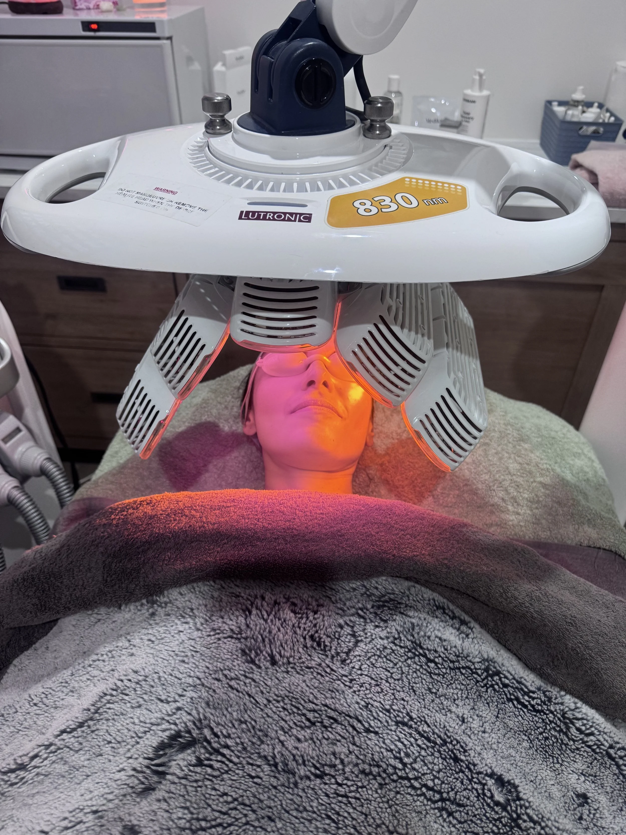 A person receiving a facial or skin treatment using a specialized machine with a face-shaped opening, labeled with '830 nm' and 'LUTRONIC', in a clinical setting. The person is lying on a towel with a towel draped over their chest.