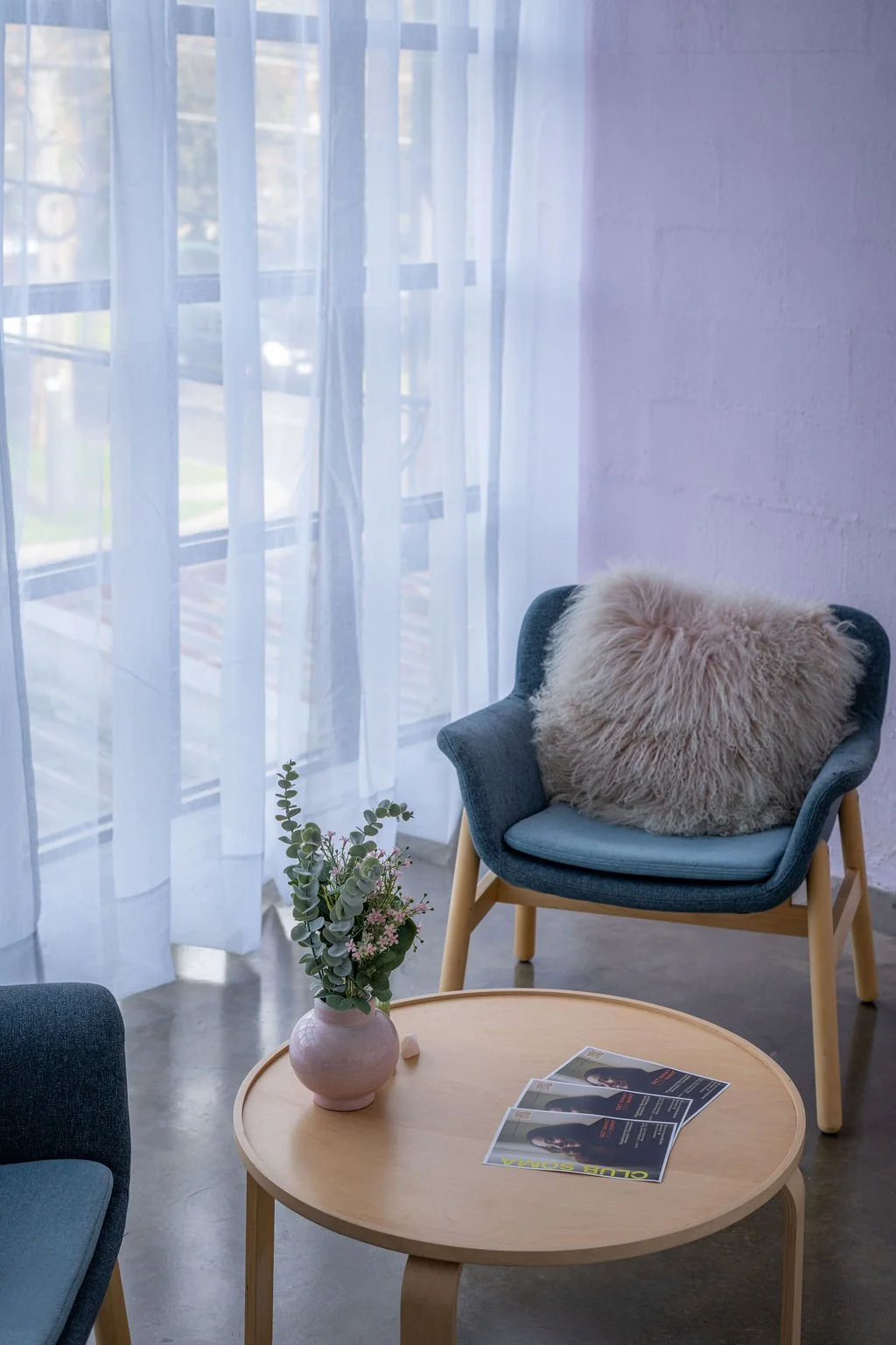 A cozy seating area with a round wooden table, a vase of greenery, brochures, and two modern armchairs with cushions next to a window with sheer curtains in a bright room.