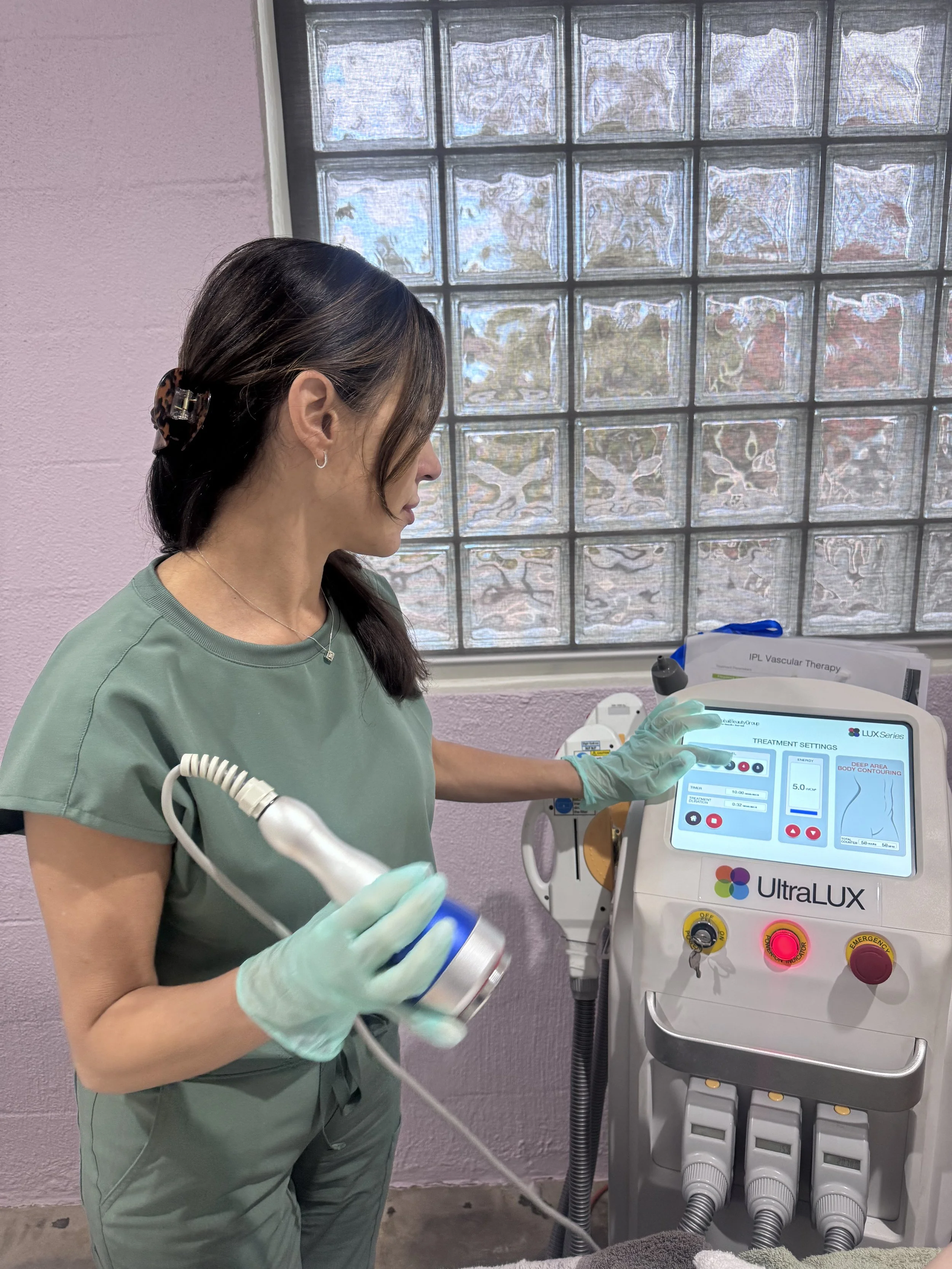 A woman in medical scrubs and gloves using a medical device labeled UltraLUX for visible light examination, standing in front of a window with glass blocks.