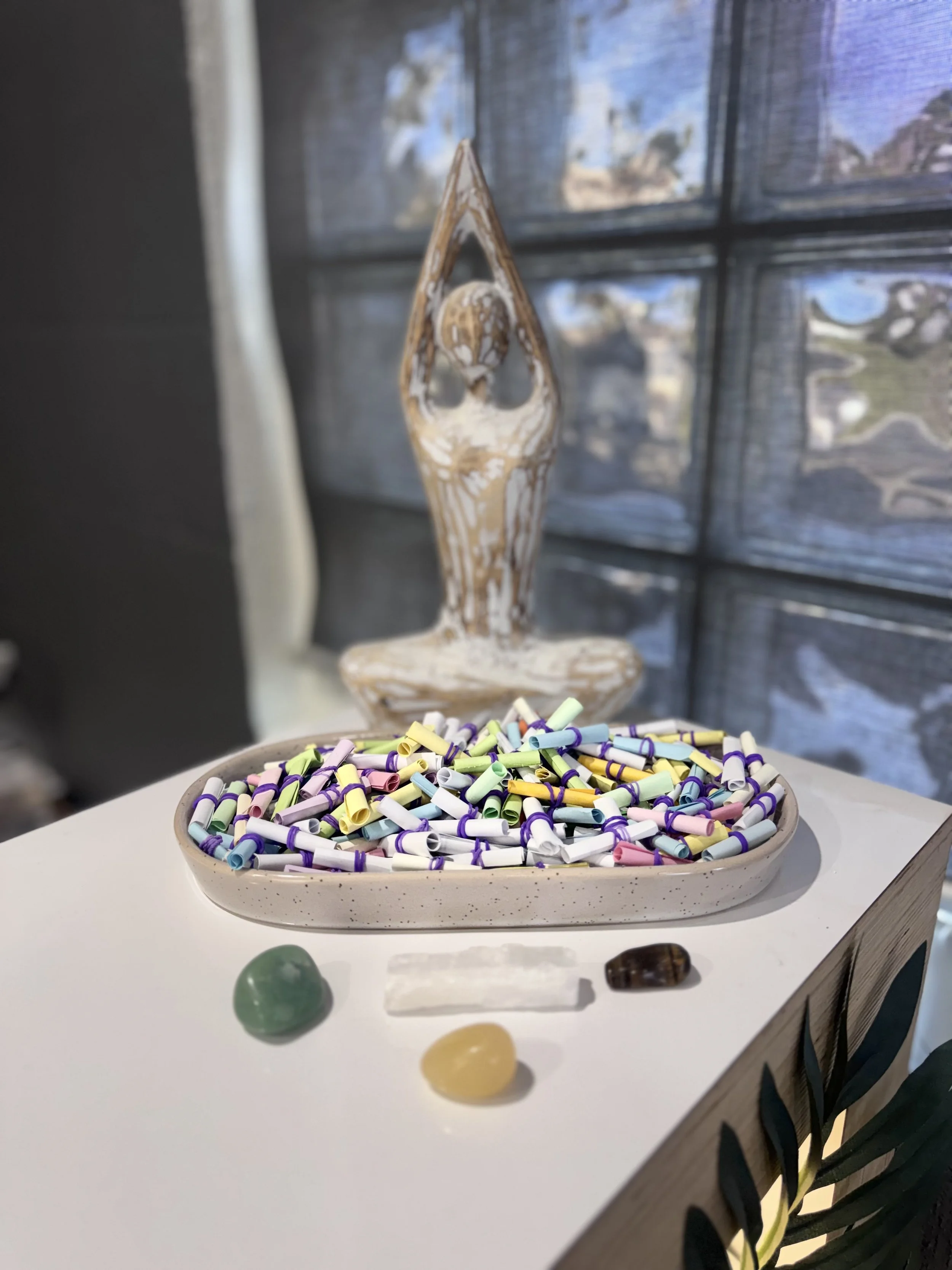 Colorful mini chalks in a ceramic bowl on a white table, with decorative stones in front and a wooden sculpture in the background.