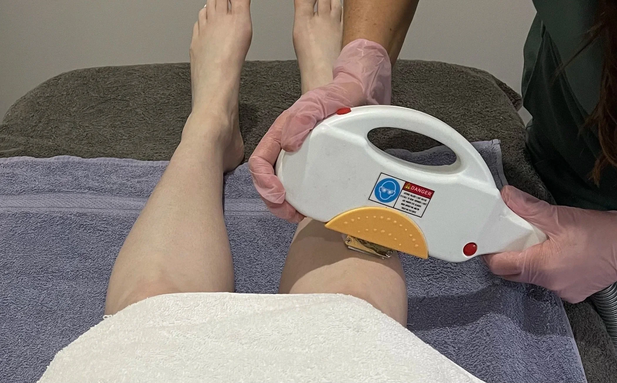 Person lying on a treatment table during a therapy session, with their legs raised. Another person, wearing pink gloves, is holding a handheld device near the person's knees. The treatment room has a brown towel covering the table and a purple towel underneath the person's legs.