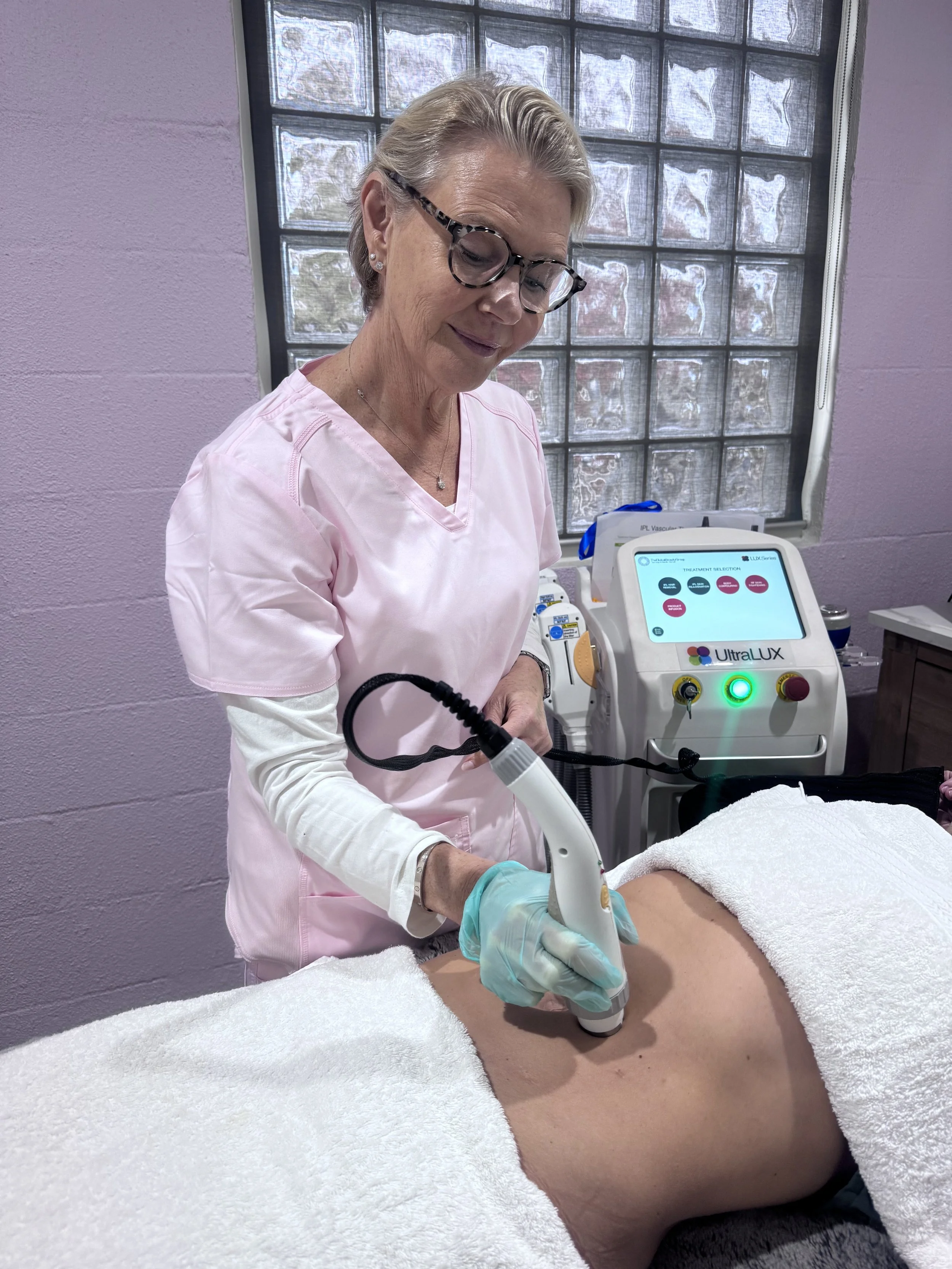 A healthcare professional, dressed in pink scrubs and wearing glasses, performs a skincare treatment on a patient lying on a treatment bed. The room has a window with glass blocks and medical equipment is visible nearby.