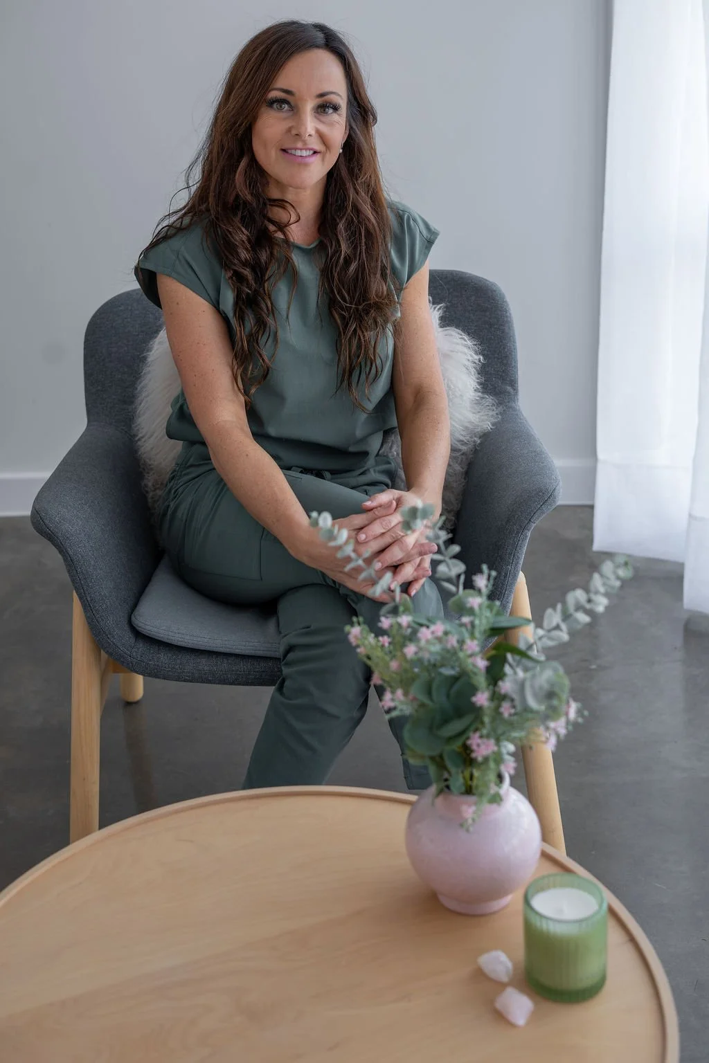 A woman with long brown hair wearing a green outfit, sitting on a gray chair with wooden legs, in a room with white walls and curtains. She is smiling and sitting with her hands clasped on her lap. Front of her, a wooden table holds a pink vase with pink and green flowers and a green candle holder with a white candle.
