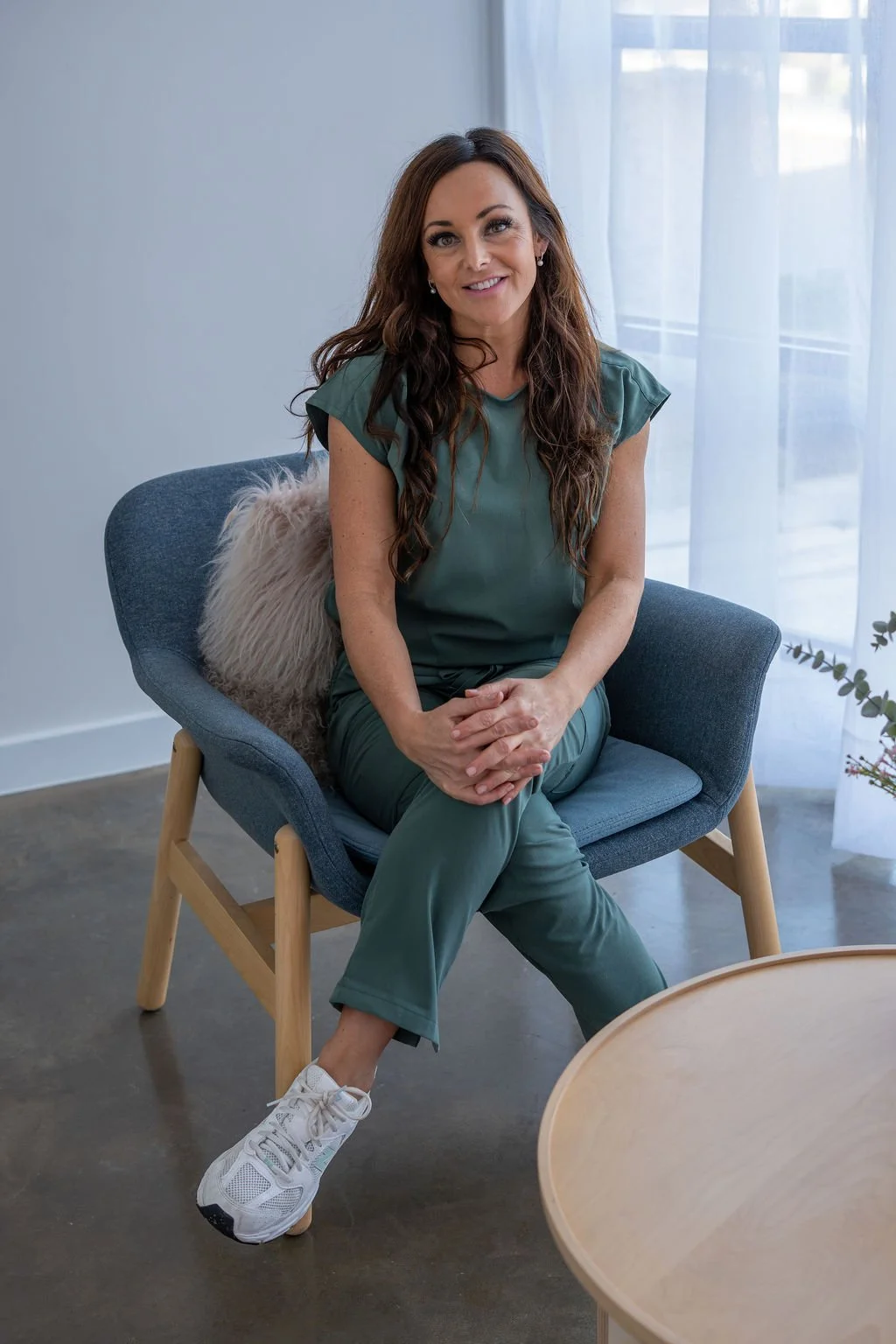 A woman sitting on a blue armchair near a window with sheer curtains, smiling and wearing green casual clothes and white sneakers, next to a fluffy pillow and a small plant.