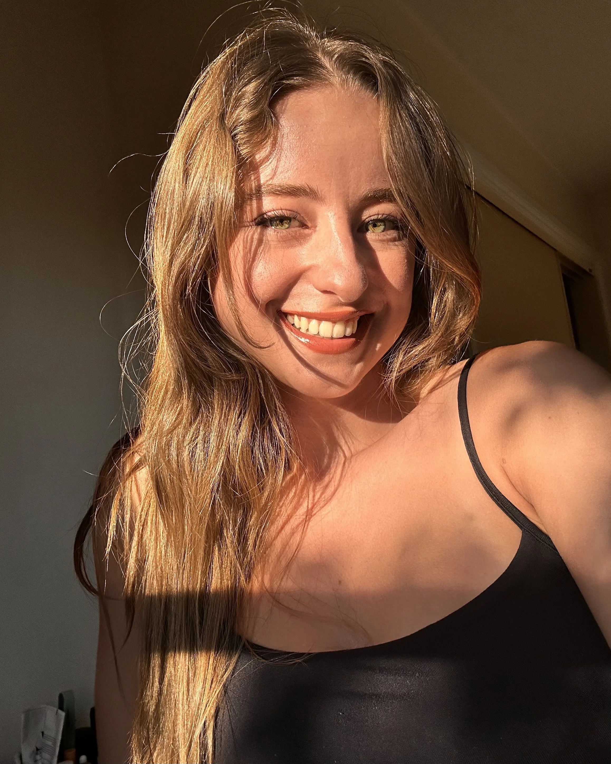 Close-up of a smiling girl with curly light brown/blonde hair, wearing a black top, in warm sunlight.