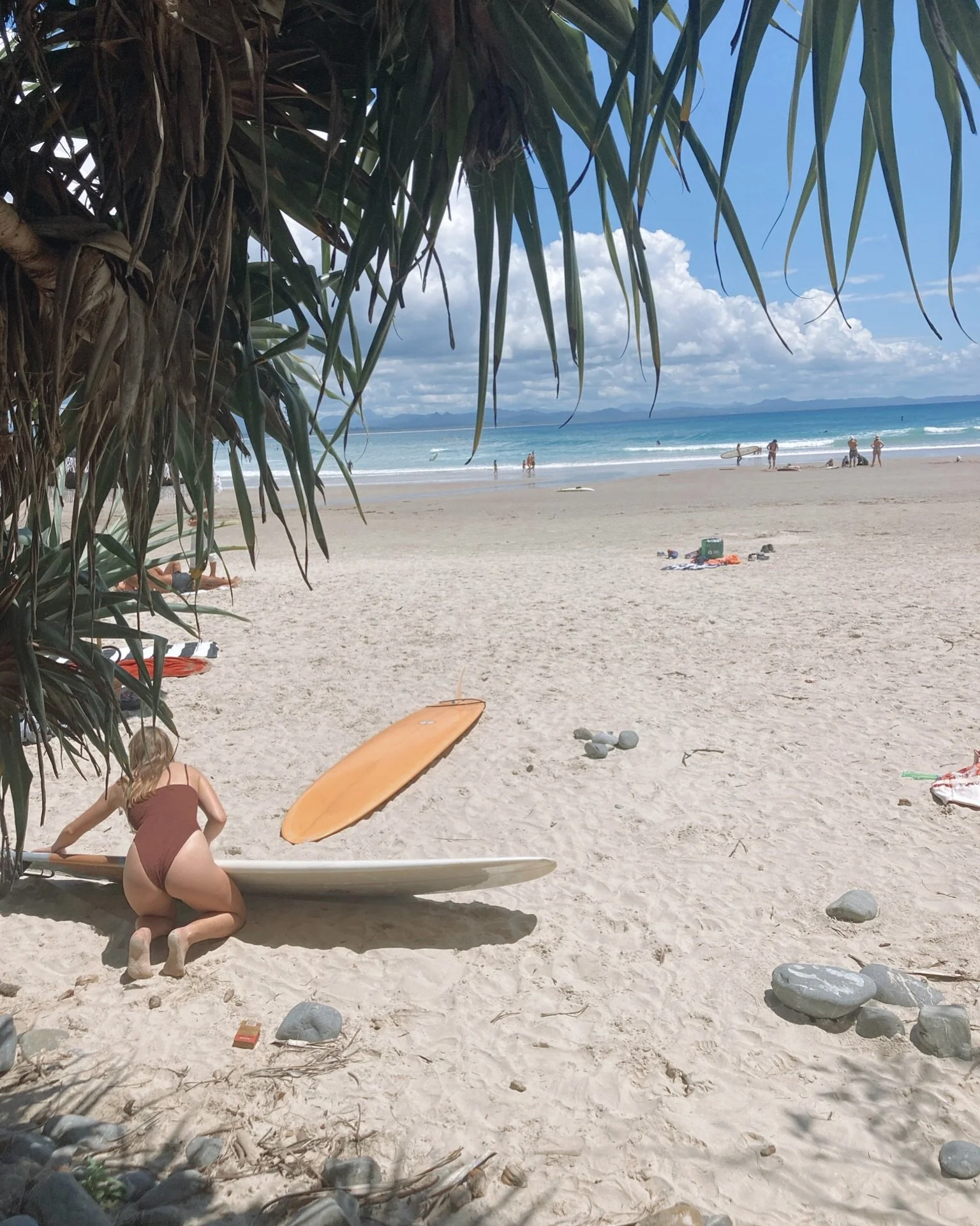 A person in a brown swimsuit crouches under a wooden paddleboard on a sandy beach, with a large leafy plant overhead. In the background, there are a few people near the shoreline, with blue ocean water and cloudy sky, and distant mountains on the horizon.