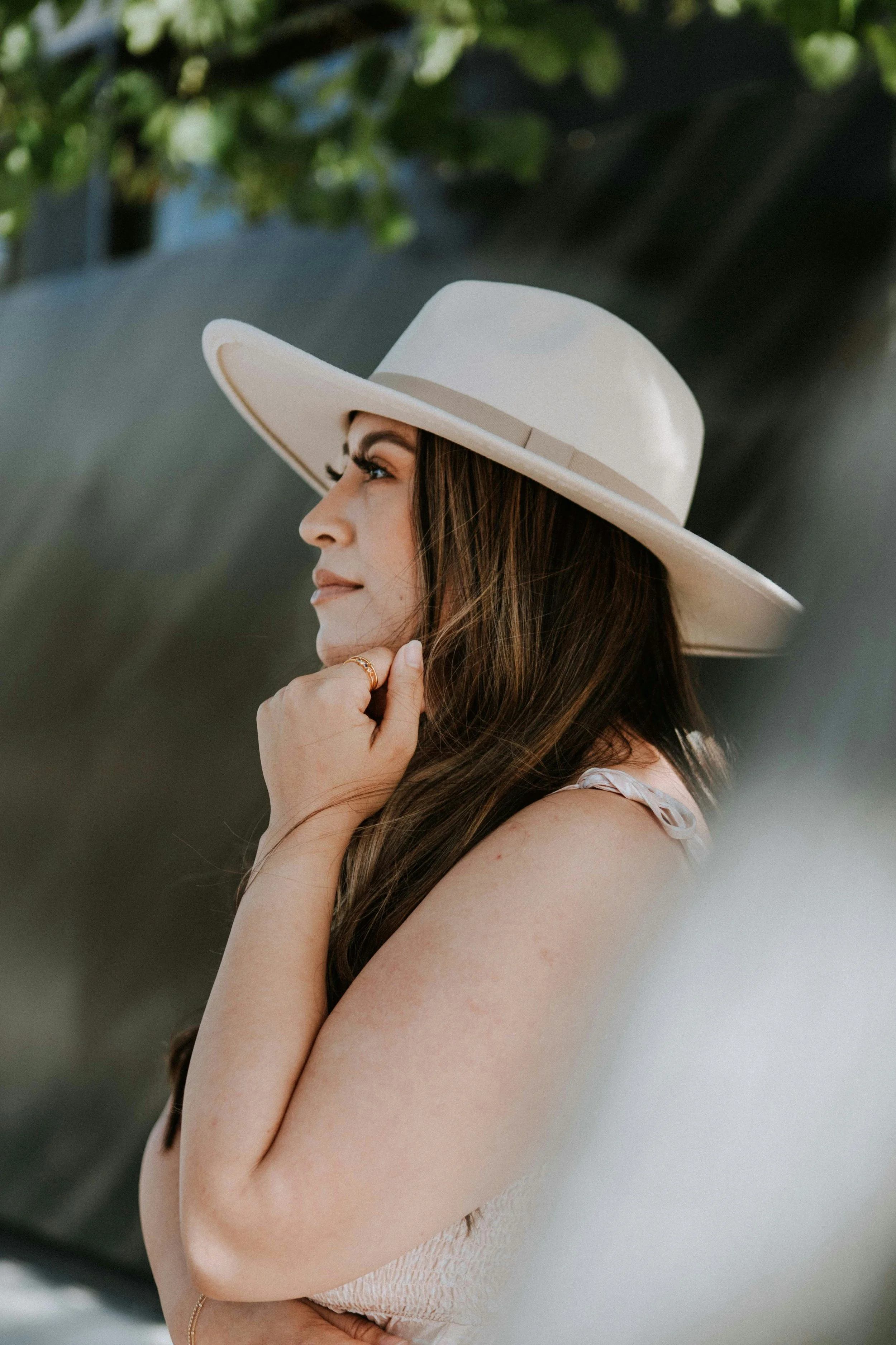 A woman with long brown hair wearing a wide-brimmed hat and a sleeveless beige top, standing outdoors with trees in the background, touching her face with her right hand.