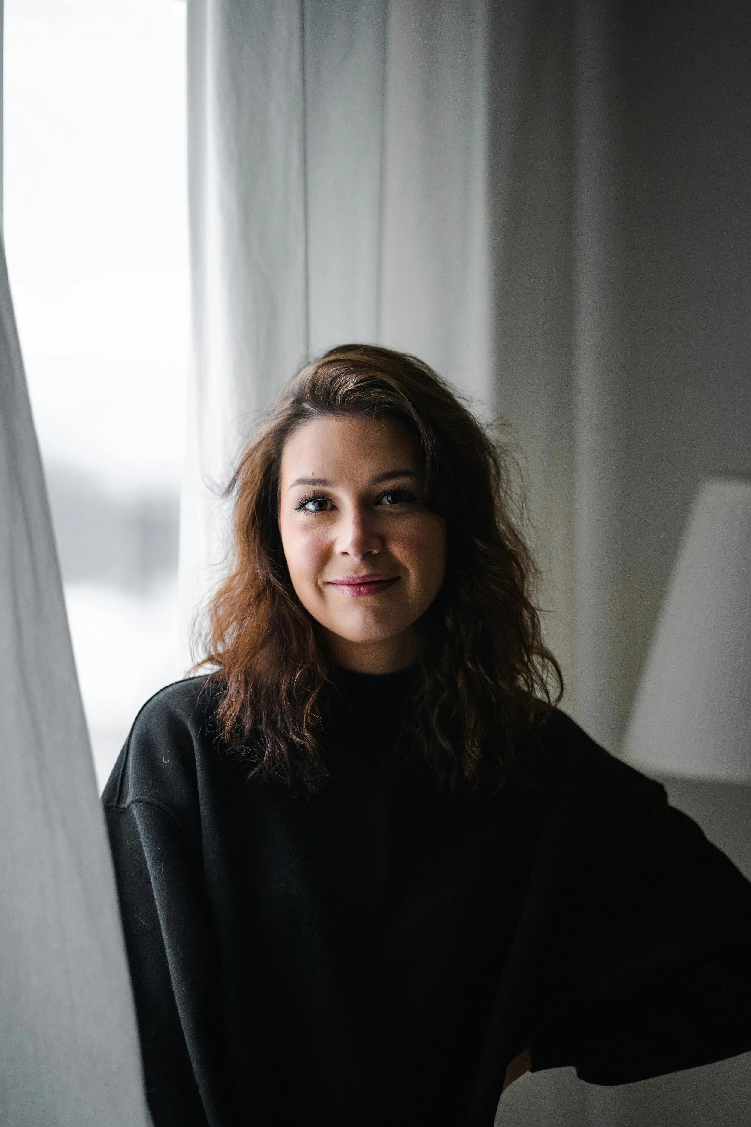 A young woman with shoulder-length brown hair wearing a black top, sitting indoors near a window with gray curtains, smiling softly.