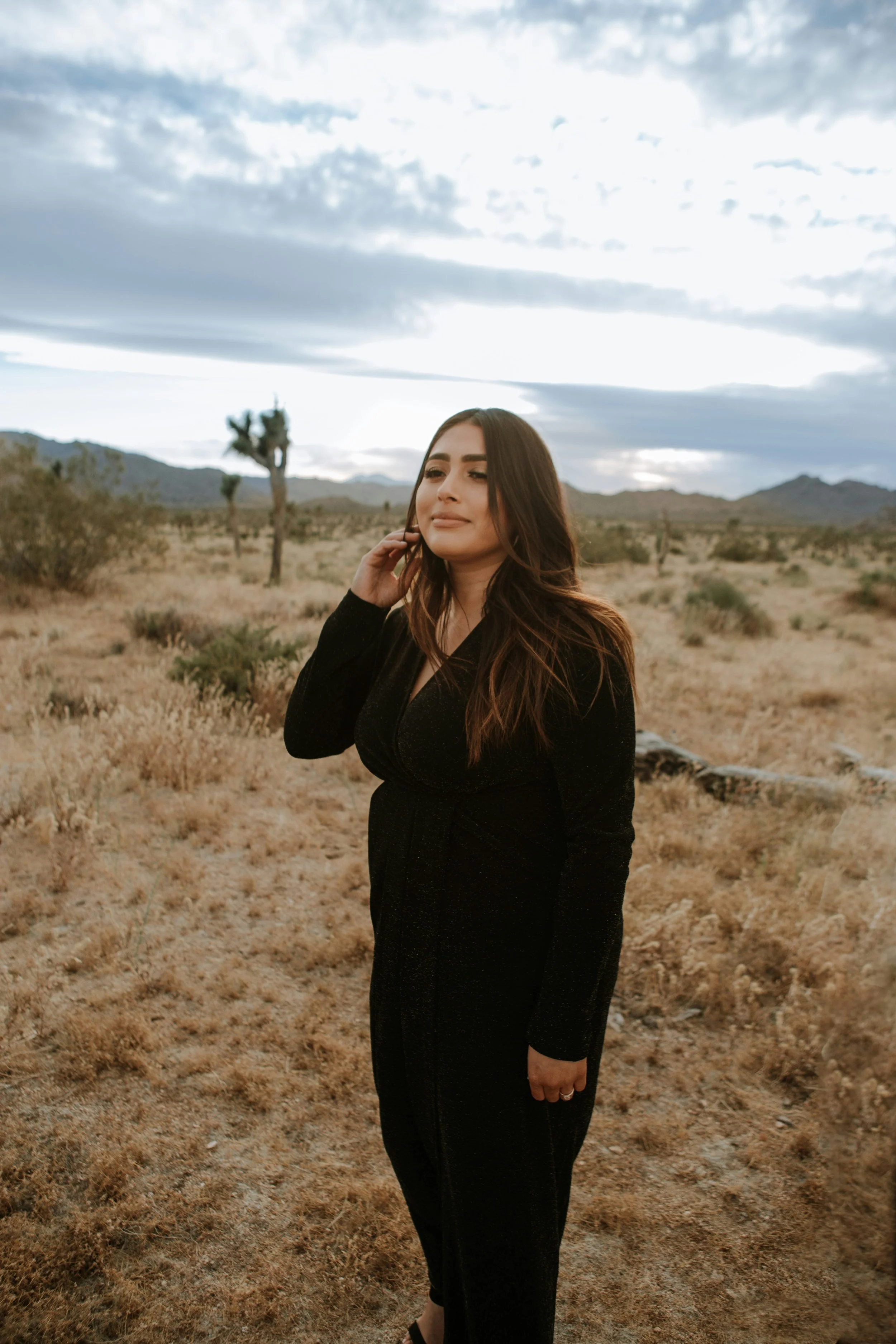 A woman with long brown hair in a black outfit standing in a desert landscape with Joshua trees and mountains in the background under a cloudy sky.