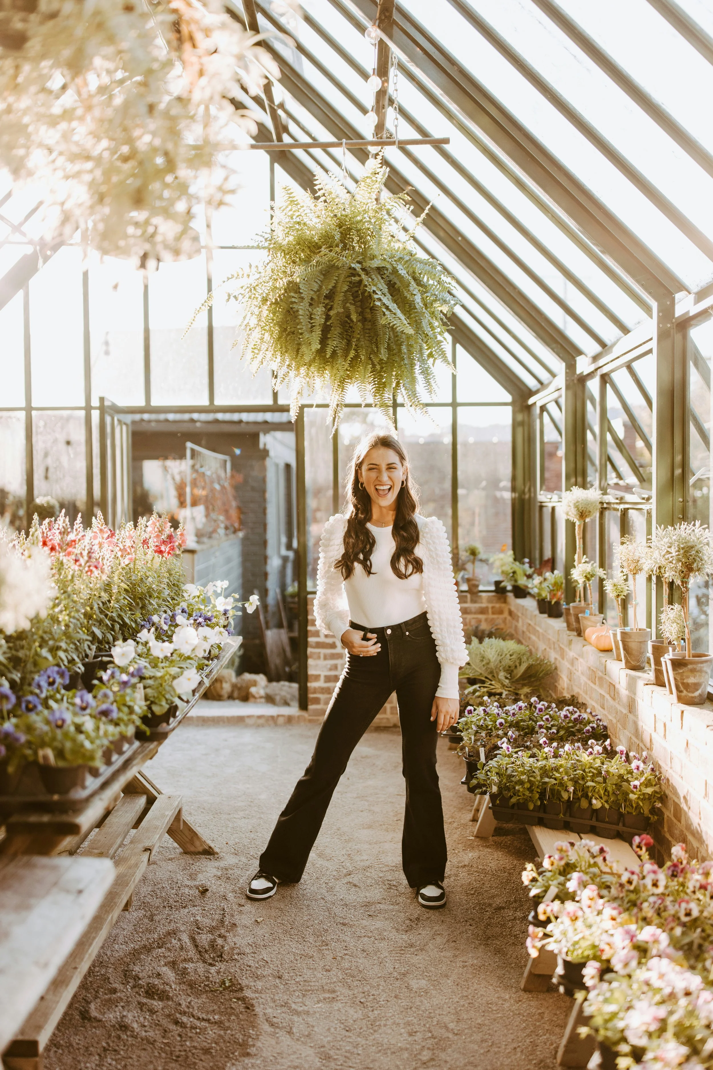 A woman with long, wavy brown hair in a greenhouse, smiling and posing with her hand on her hip, surrounded by colorful potted flowers and plants, with large glass windows and hanging plants in the background.