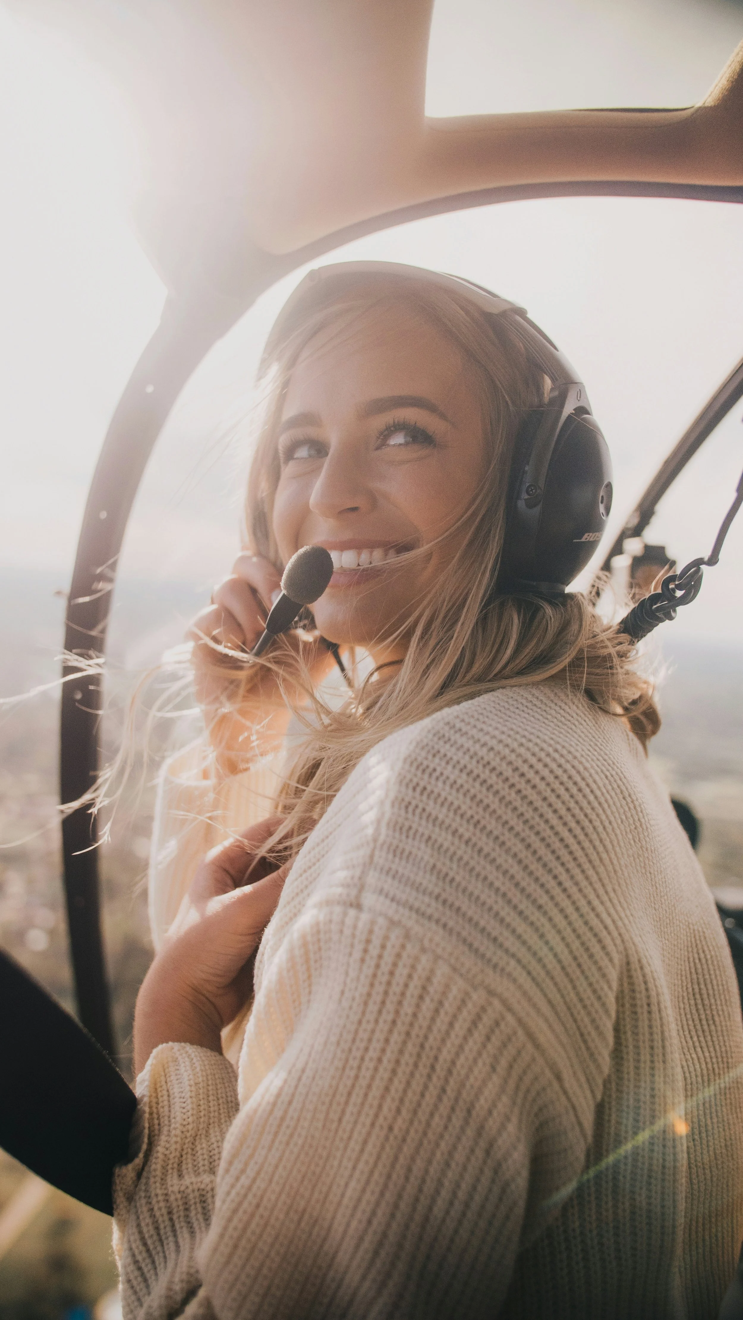 A woman with blonde hair smiling and looking up, wearing a headset with a microphone inside a helicopter.