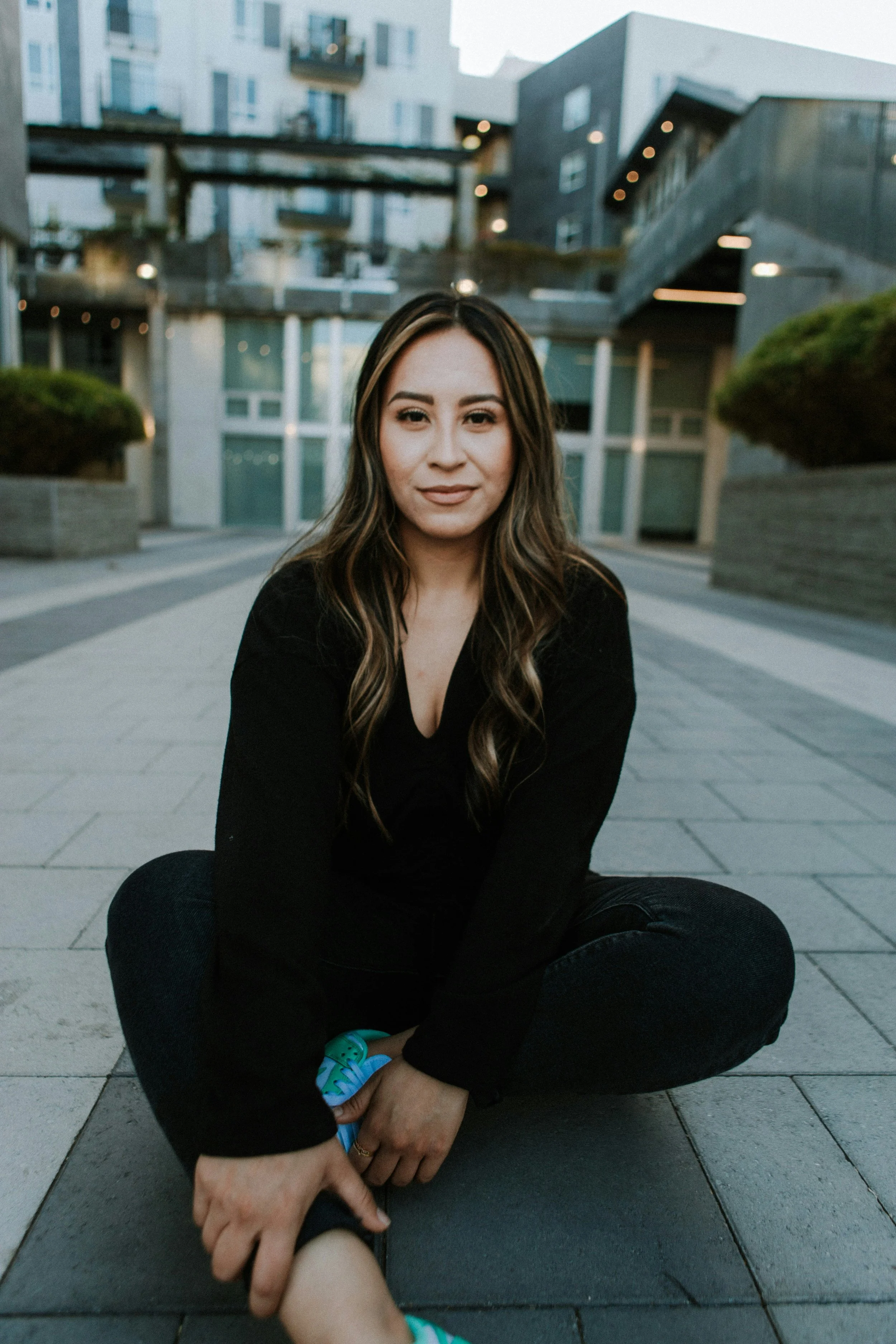 A woman with long wavy hair, wearing a black top, sitting cross-legged on the pavement outdoors, with modern buildings in the background.