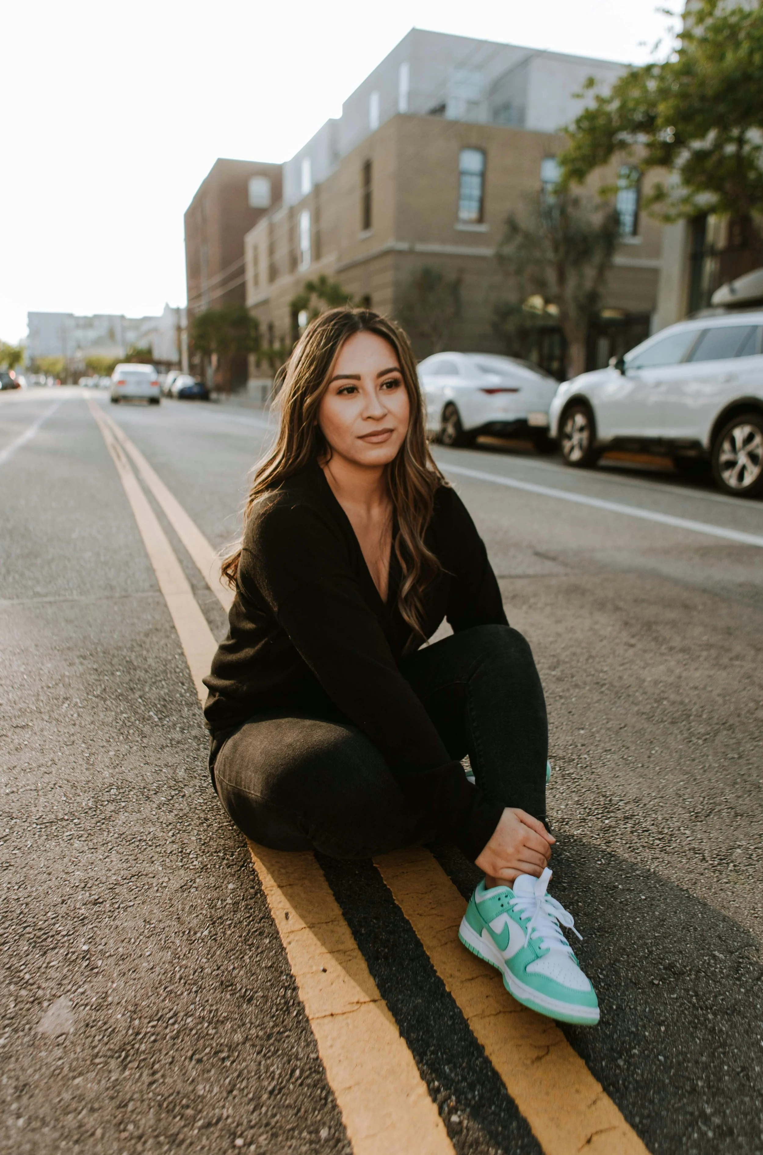 A young woman sitting on a city street with her legs crossed, wearing a black long sleeve shirt, black pants, and white and green sneakers. She has long, wavy brown hair and is looking into the distance. Parked cars and buildings are visible in the background, and the sun is setting. The street has double yellow lines and asphalt.
