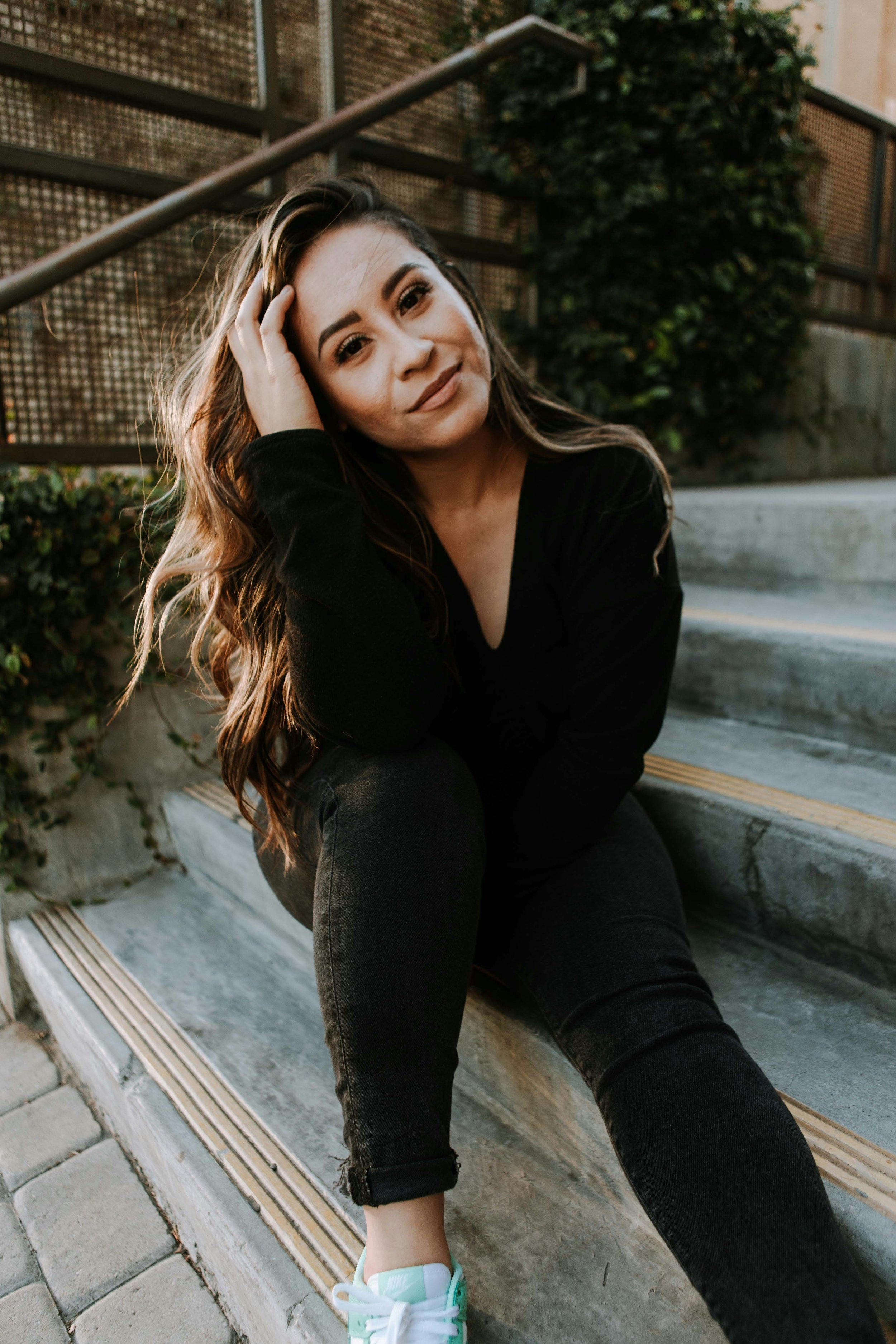 A young woman with long brown hair, wearing a black sweater and black pants, sitting on outdoor steps with a relaxed expression, touching her hair with one hand. She is wearing white and mint green sneakers. There is greenery and a metal fence in the background.