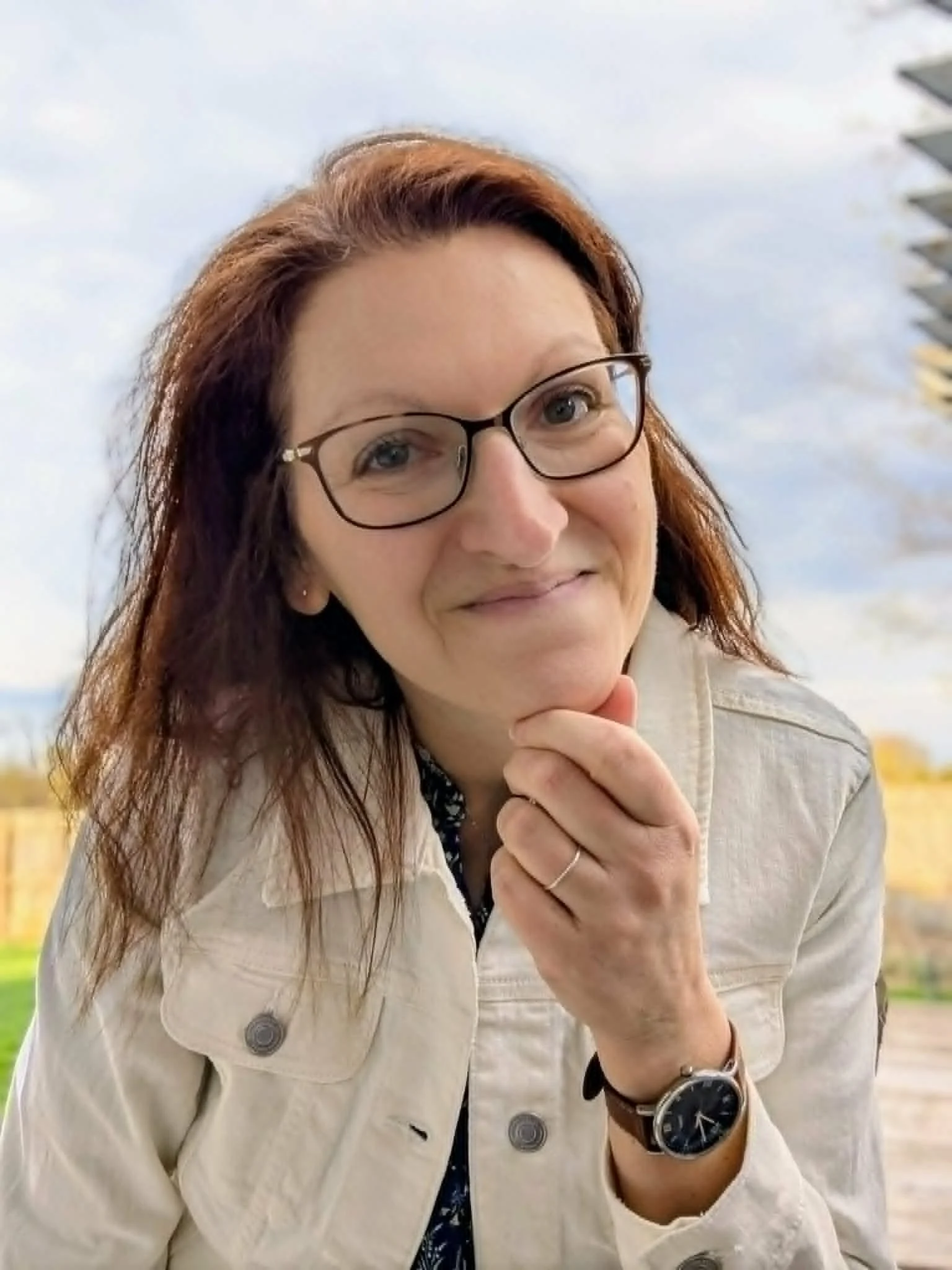 Close-up of a woman wearing glasses, smiling, with her hand resting on her chin, outdoors with a blue sky and clouds in the background.