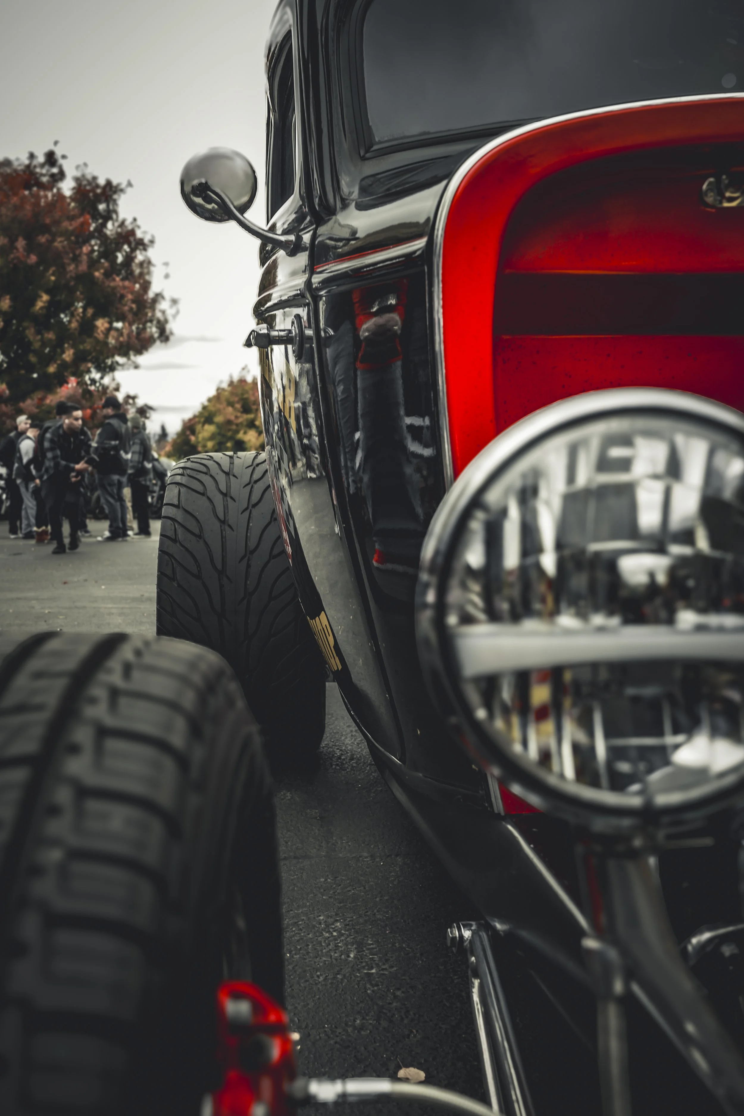 Close-up of a shiny black vehicle, possibly a scooter or motorcycle, with a red interior and large tires, parked on a street. In the background, a group of people are standing outdoors, some looking at their phones, with trees showing fall foliage.